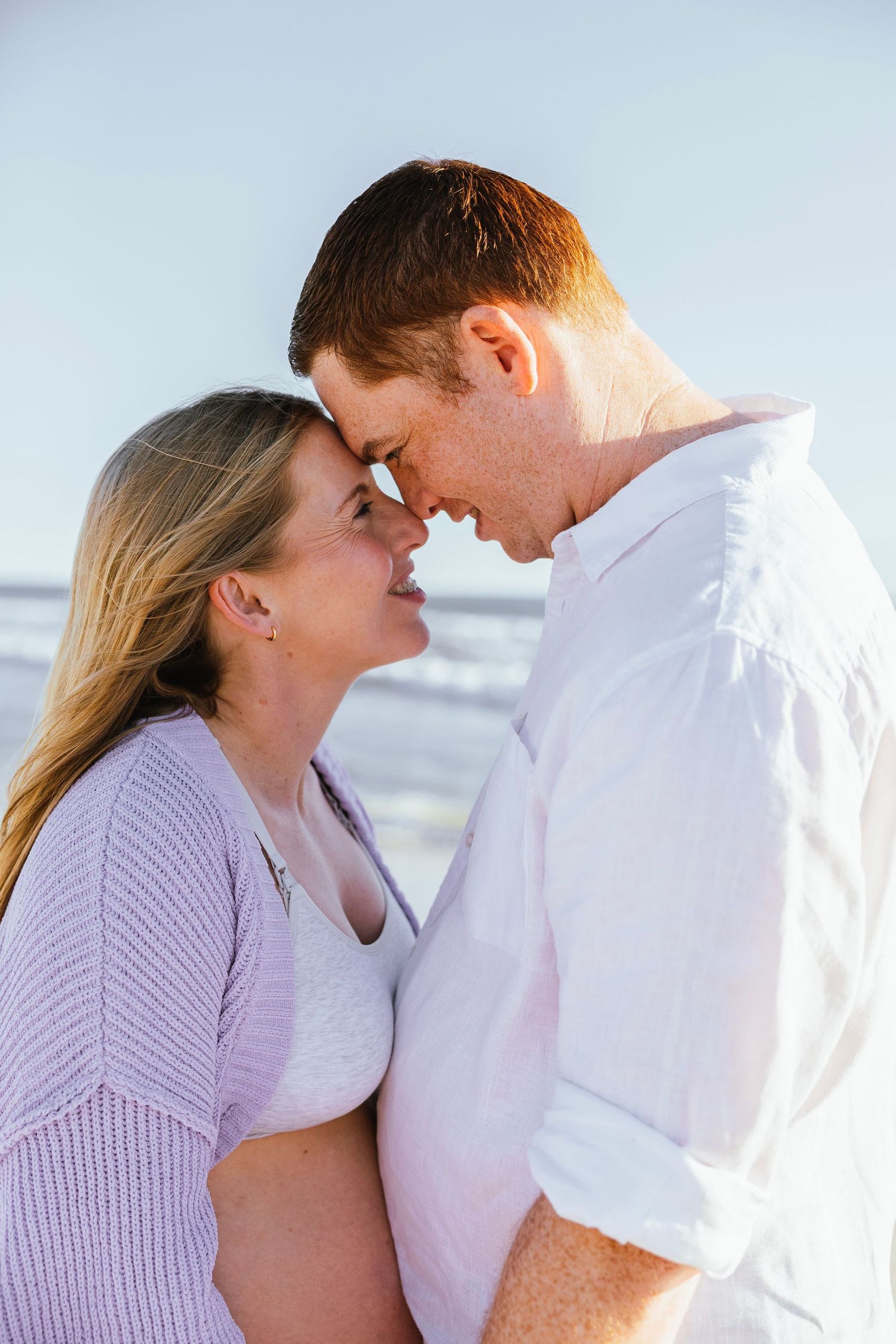 A romantic couple touches foreheads and smiles tenderly on a bright beach, the woman in lavender and man in white shirt.