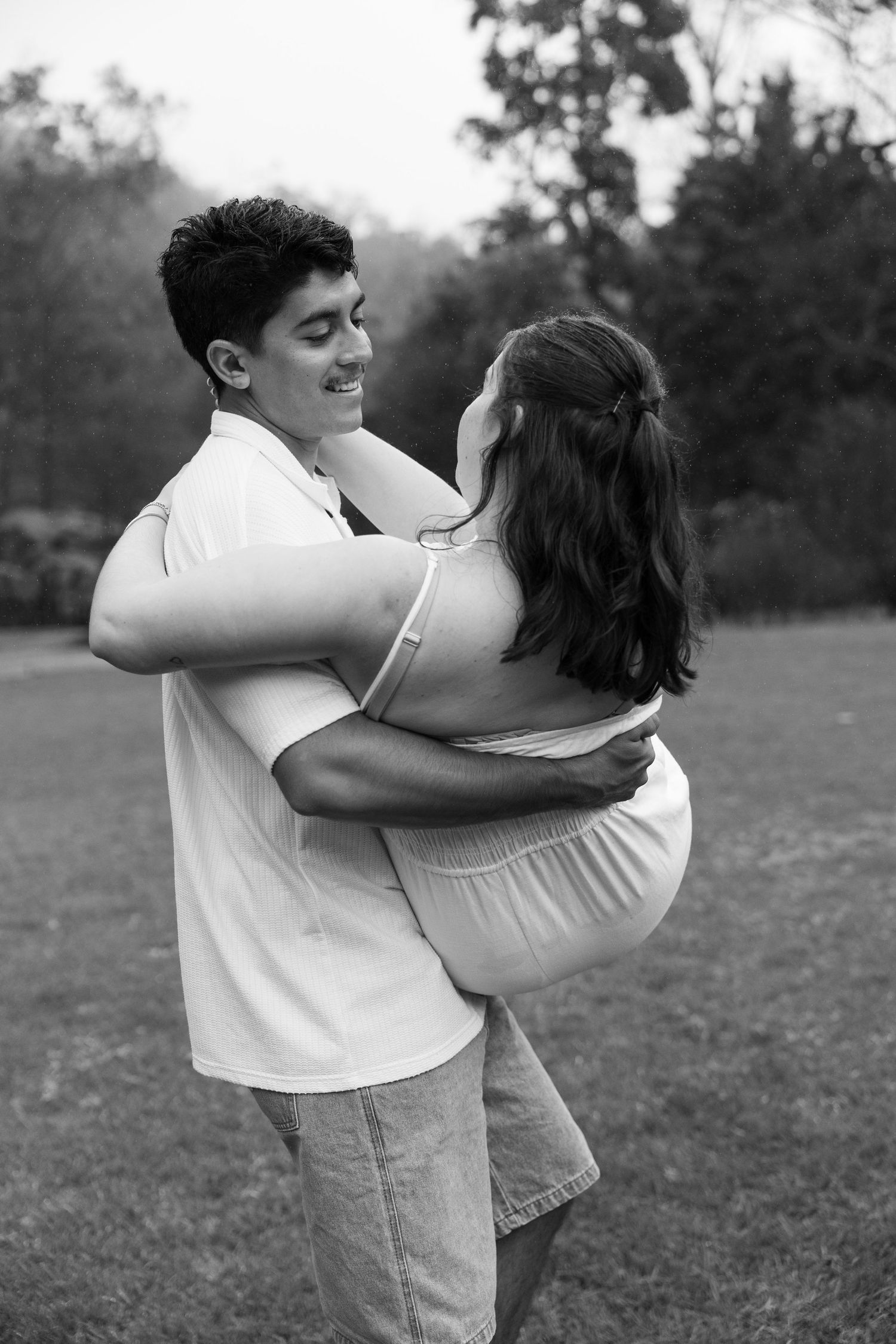Black and white photo of a young couple embracing and smiling in a lush outdoor park setting.