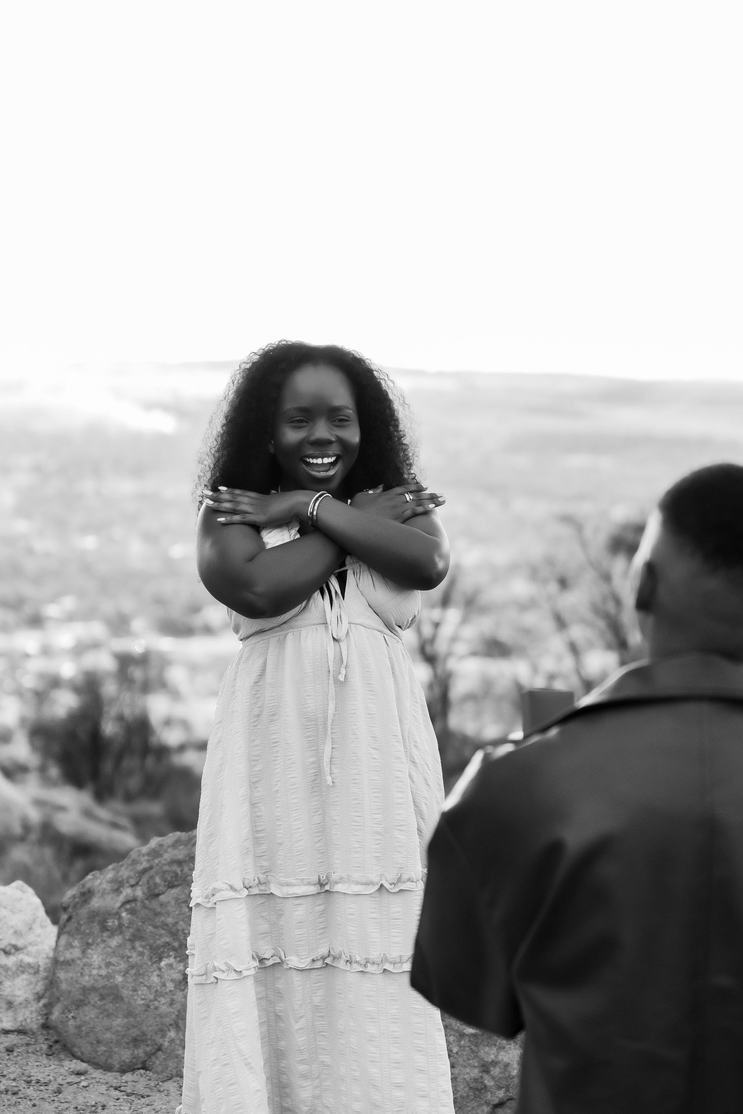 Black and white photo of a smiling young woman in a white dress with arms crossed at an outdoor overlook.