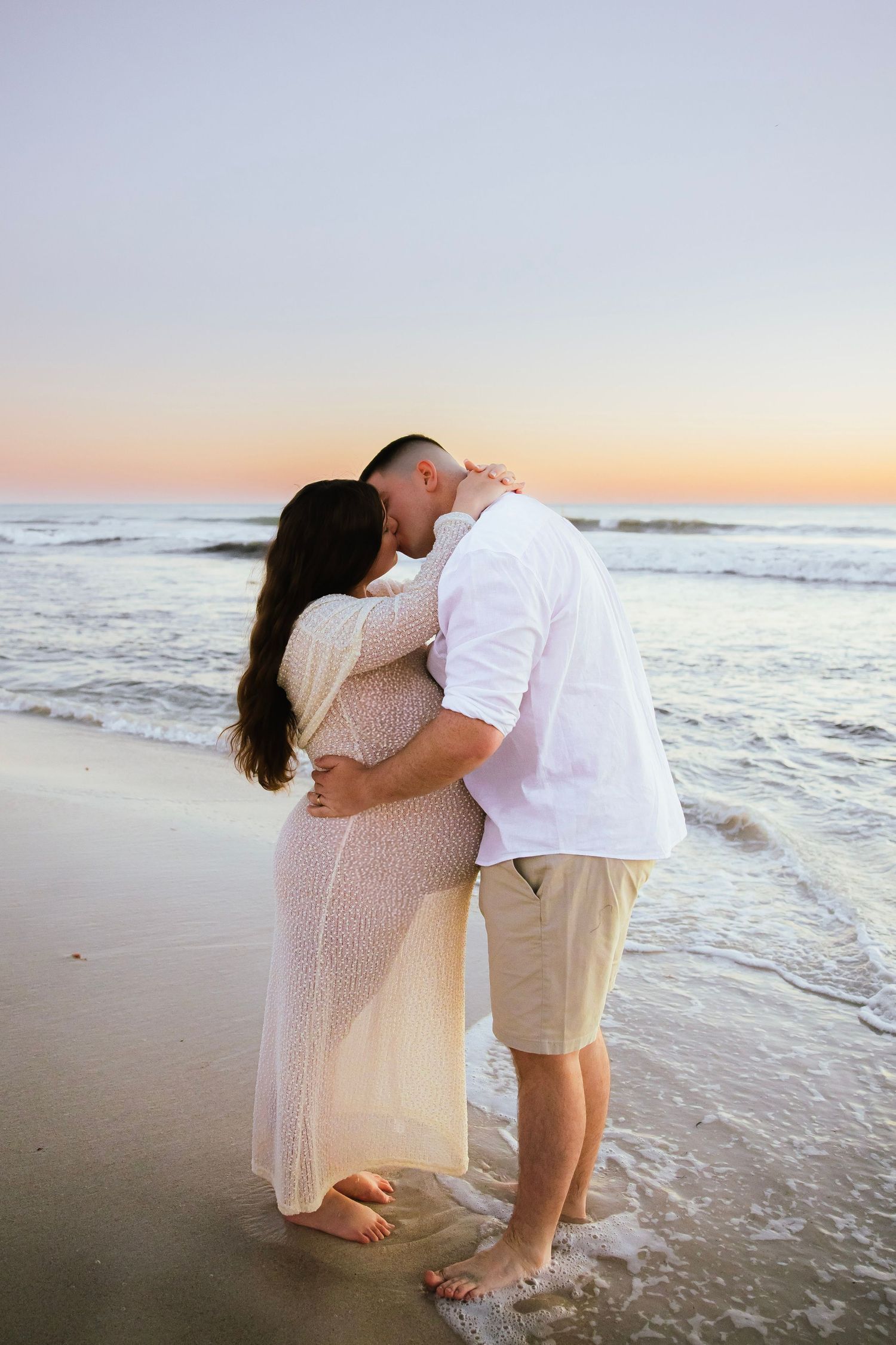 Couple sharing a kiss on sandy beach at sunset, pregnant woman in white lace maternity dress.