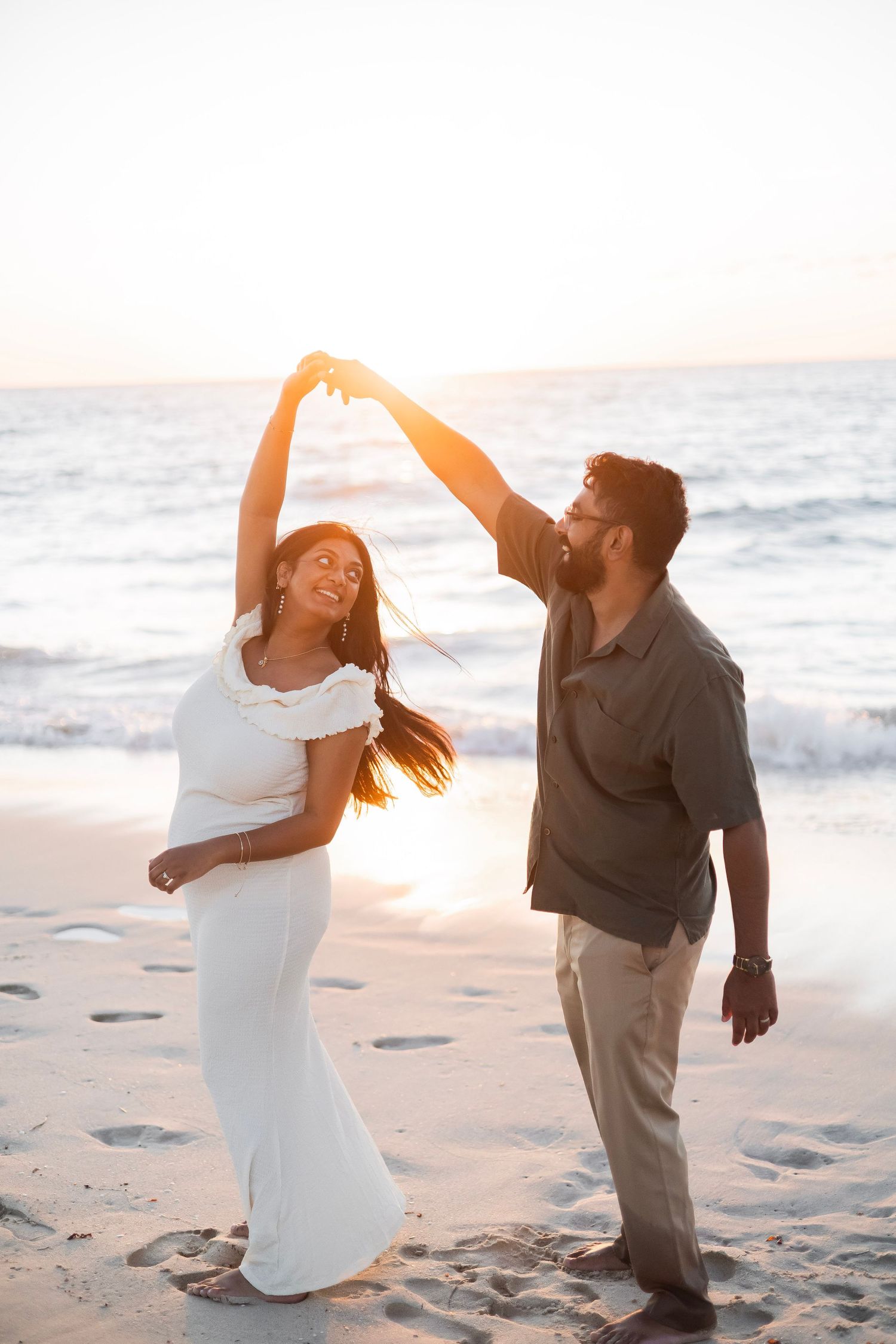 Couple dancing on beach at sunset, woman in white dress being twirled by man in olive shirt.