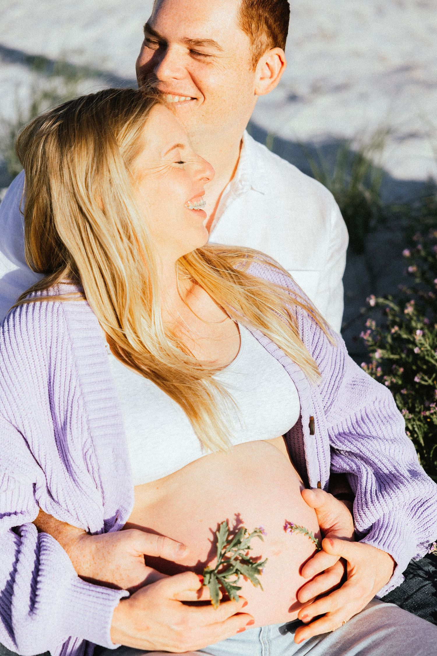 Happy couple sitting outdoors, pregnant woman cradling baby bump with flowers, partner embracing her from behind in warm sunlight.