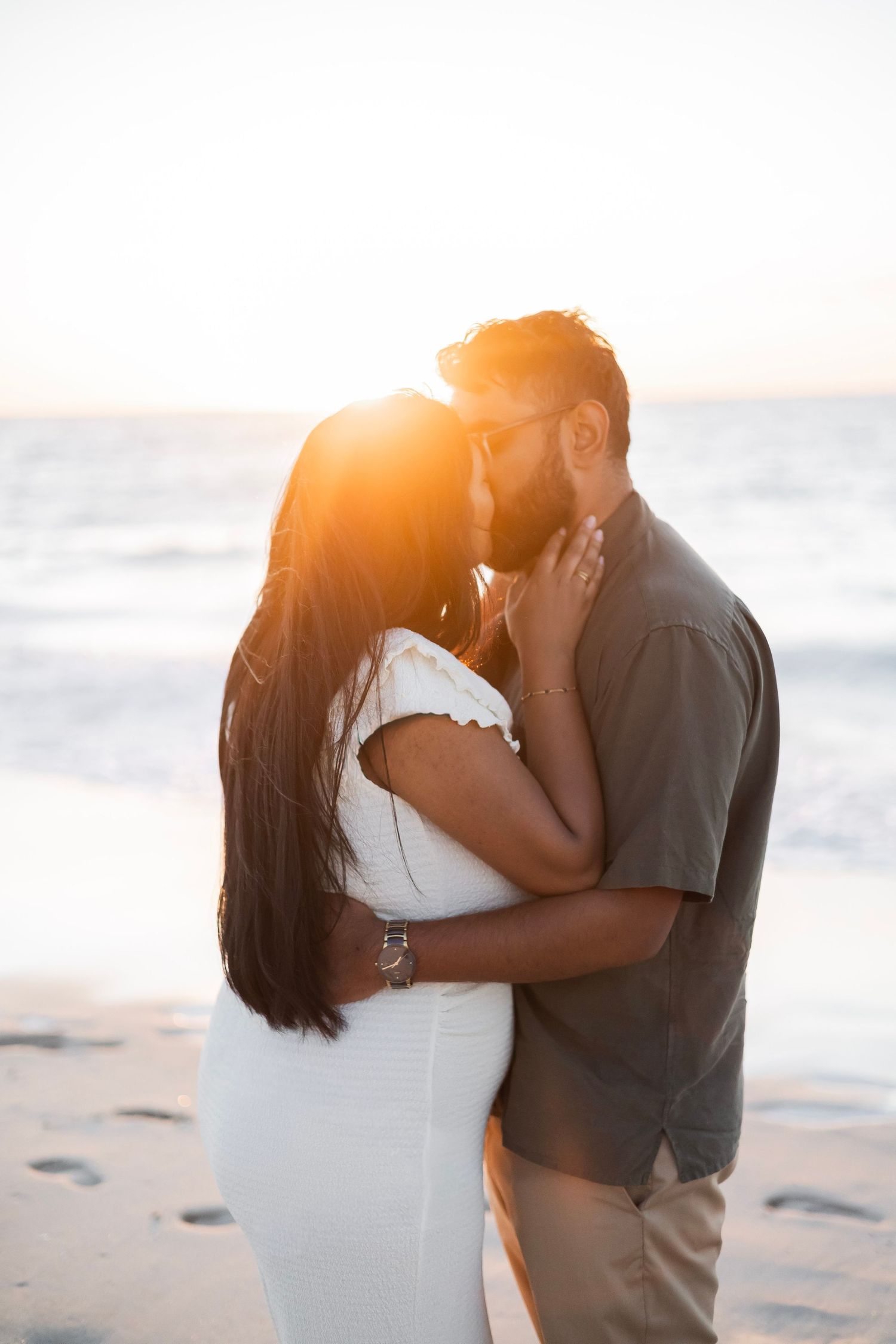 Couple sharing a romantic kiss on the beach at golden sunset, woman in white dress embraced by bearded man in gray shirt.