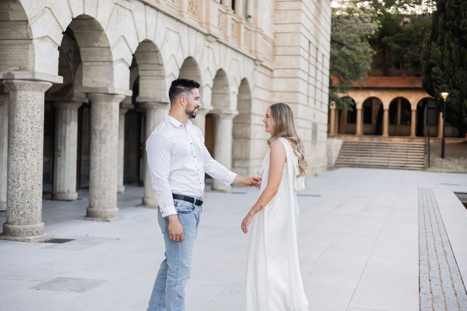 Couple holding hands and smiling at each other in front of an elegant arched stone building.