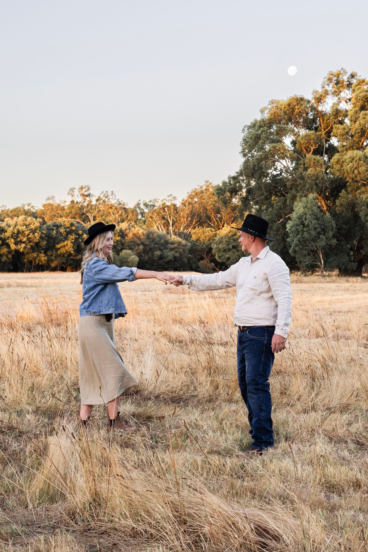 Man and woman in cowboy hats holding hands in a dry golden field with full moon rising.