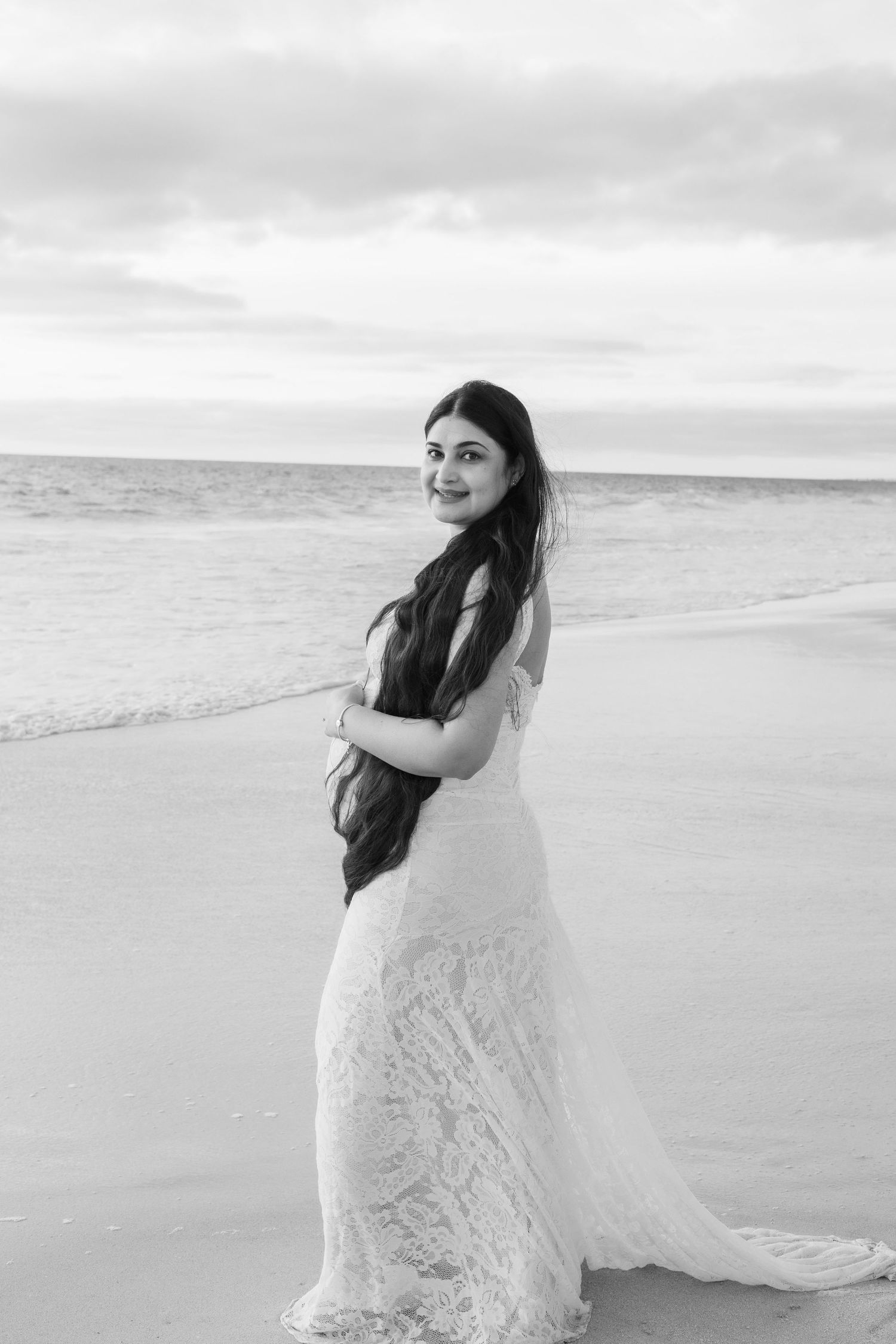 Smiling bride in white lace gown with long hair posing on beach at sunset, black and white portrait.
