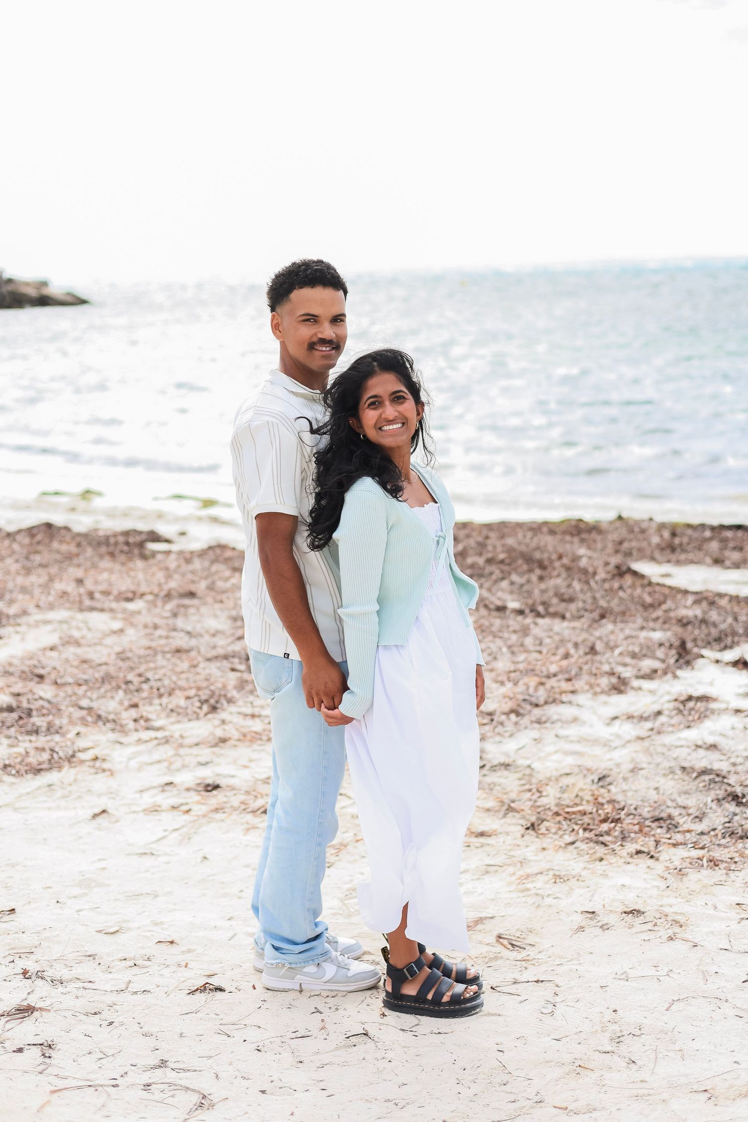 A smiling couple poses together holding hands on a sandy beach with the ocean in the background.