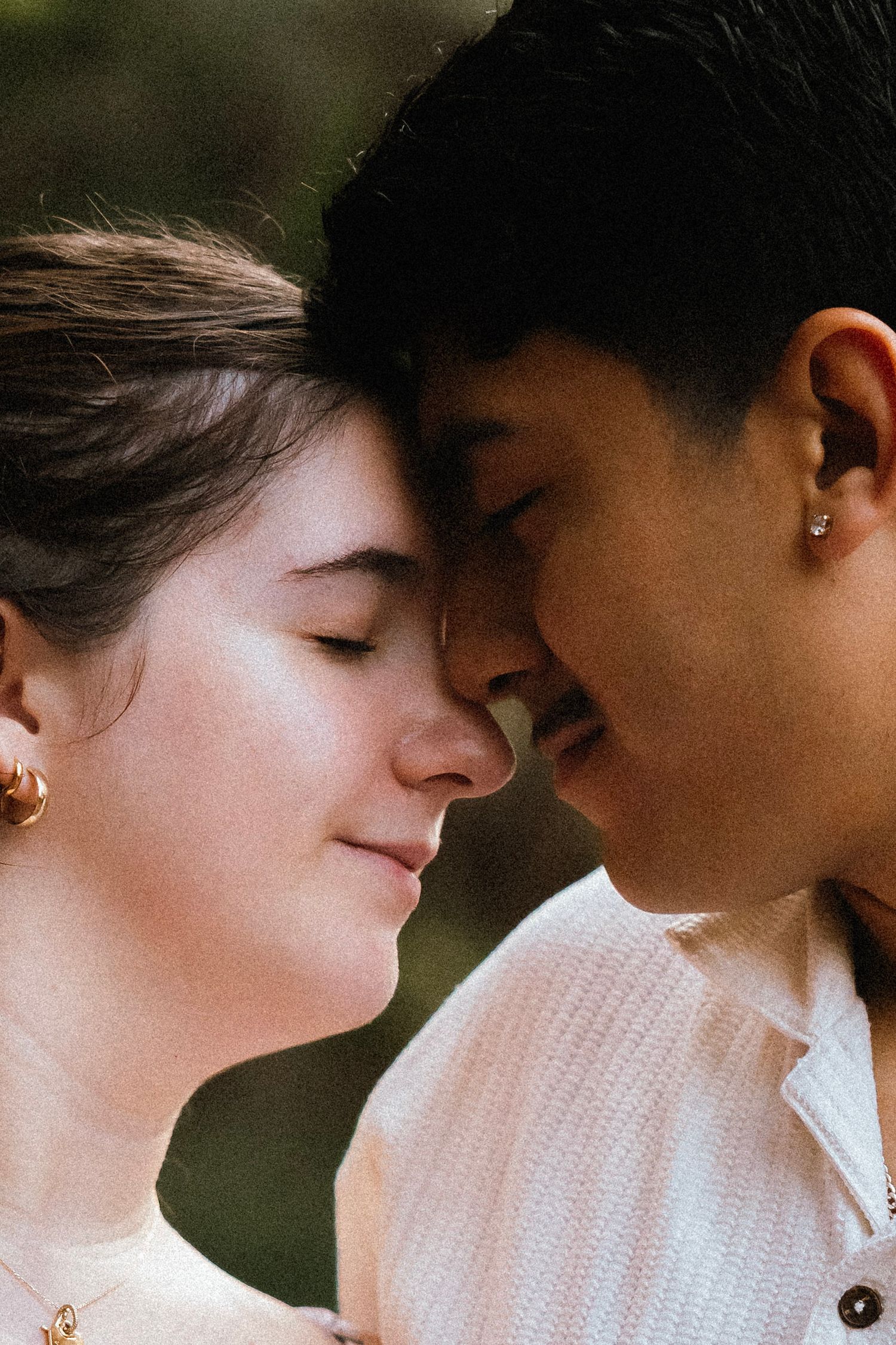 A couple shares an intimate forehead-to-forehead moment, eyes closed, both wearing light-colored tops outdoors.