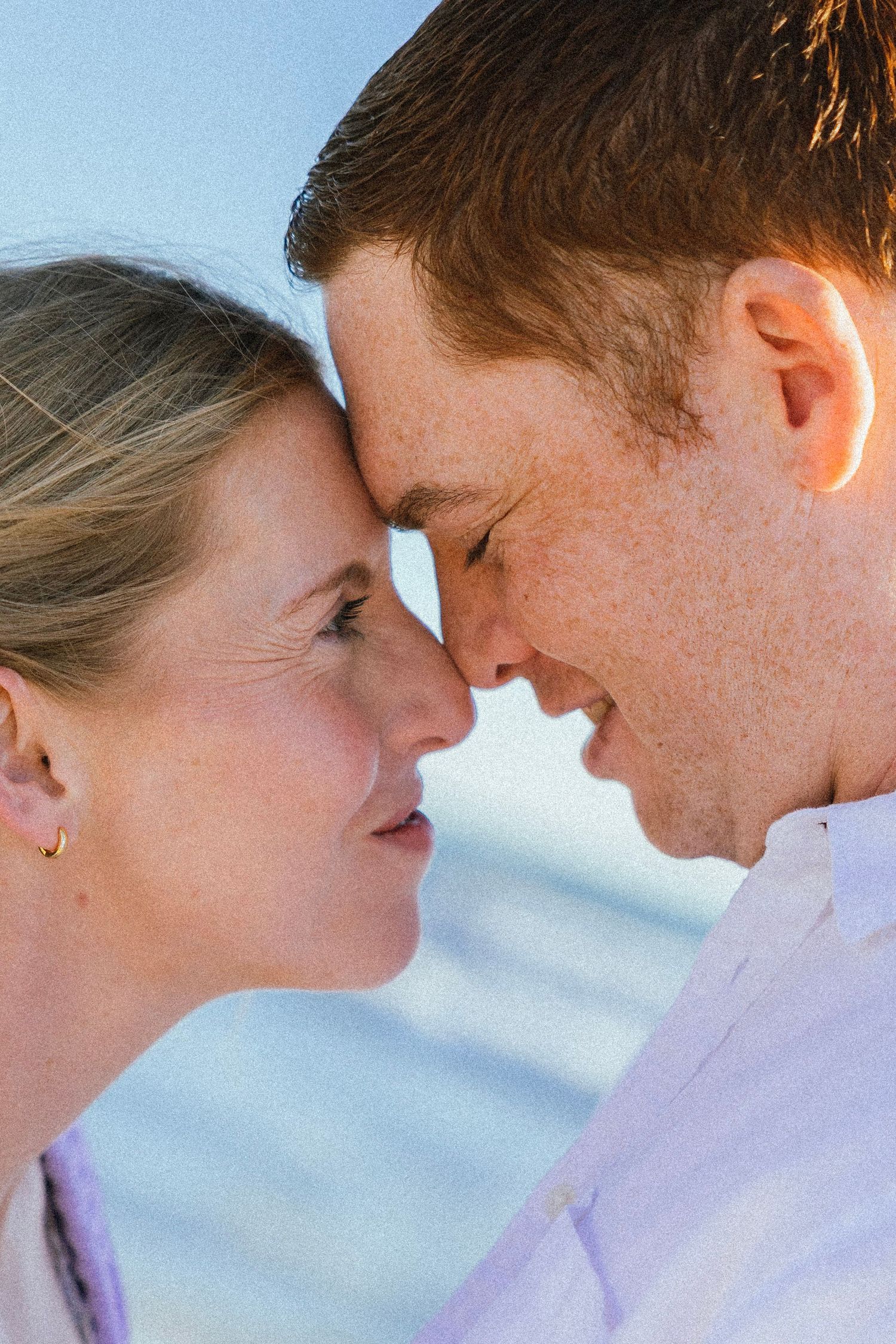 Close-up of smiling couple touching foreheads tenderly against a soft blue sky background.