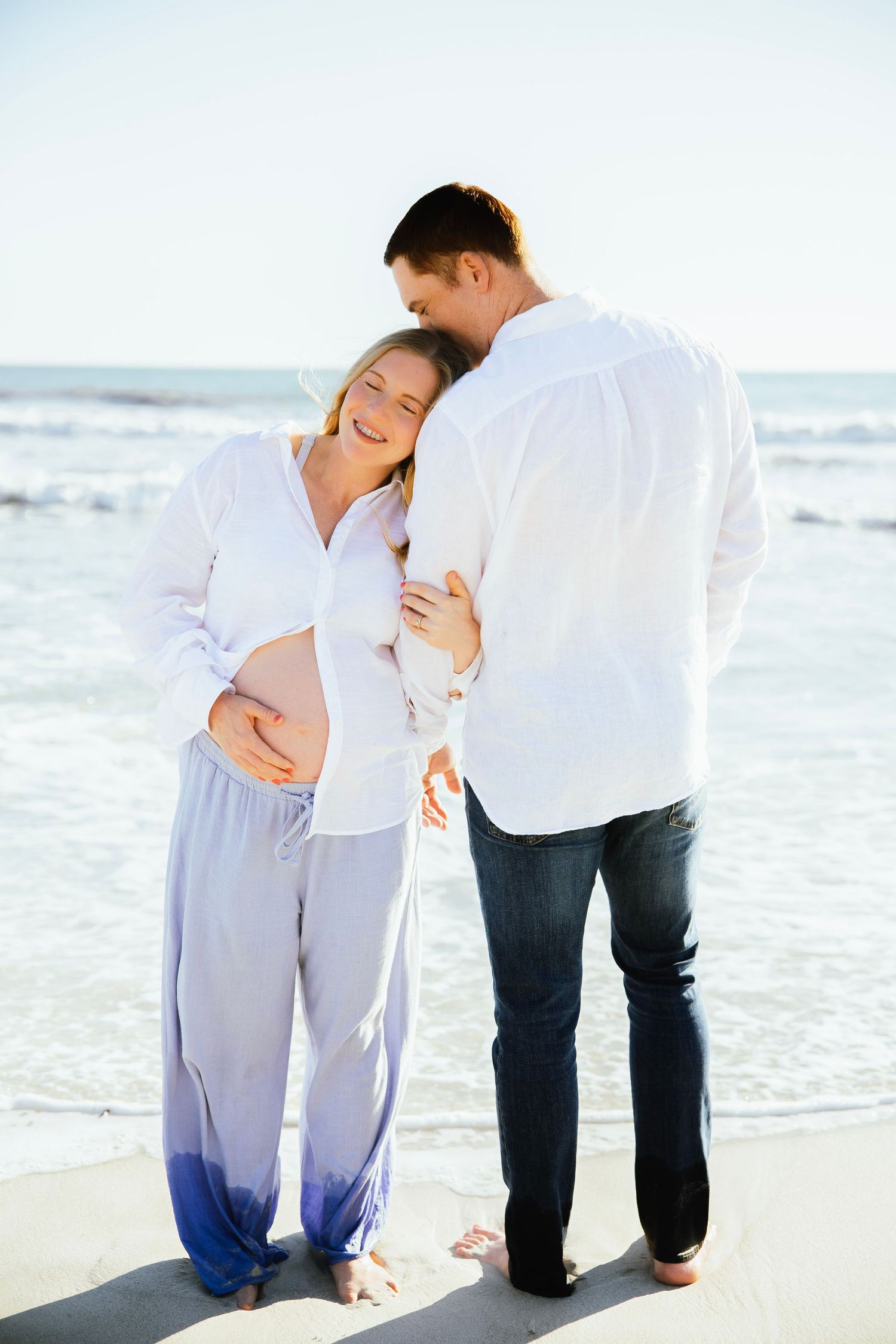 Pregnant couple embracing on beach, woman in white shirt and blue pants, man in white shirt and jeans, ocean backdrop.