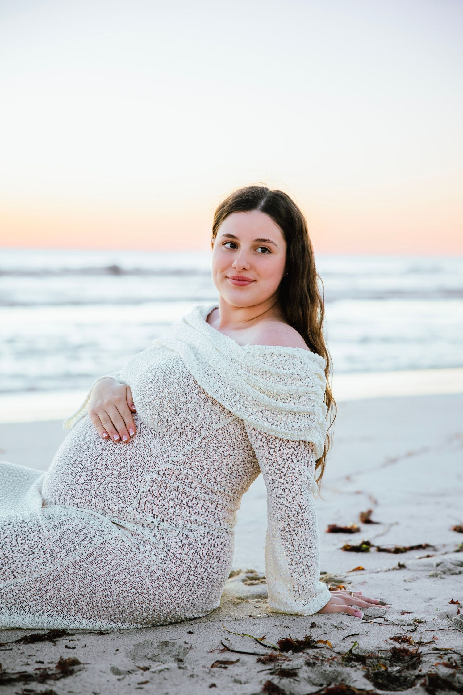 Pregnant woman in white knit dress sitting on sandy beach at sunset, smiling at camera.