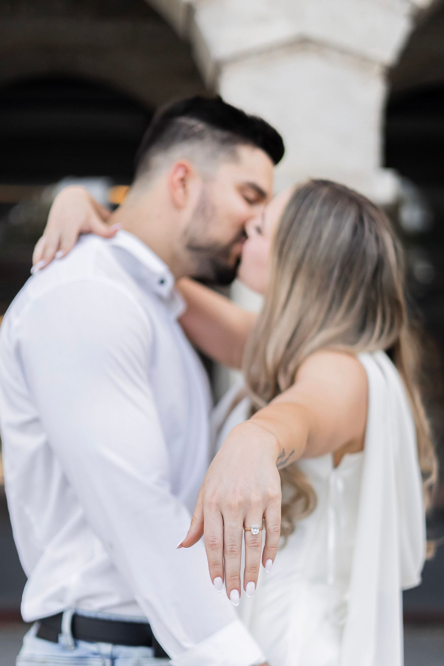 Couple kissing while woman displays diamond engagement ring on outstretched hand toward camera.