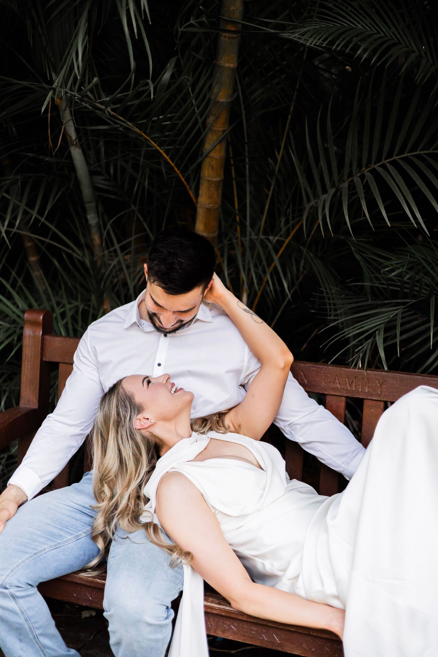 Woman lying on a wooden bench gazing up at her partner surrounded by lush tropical palm greenery.