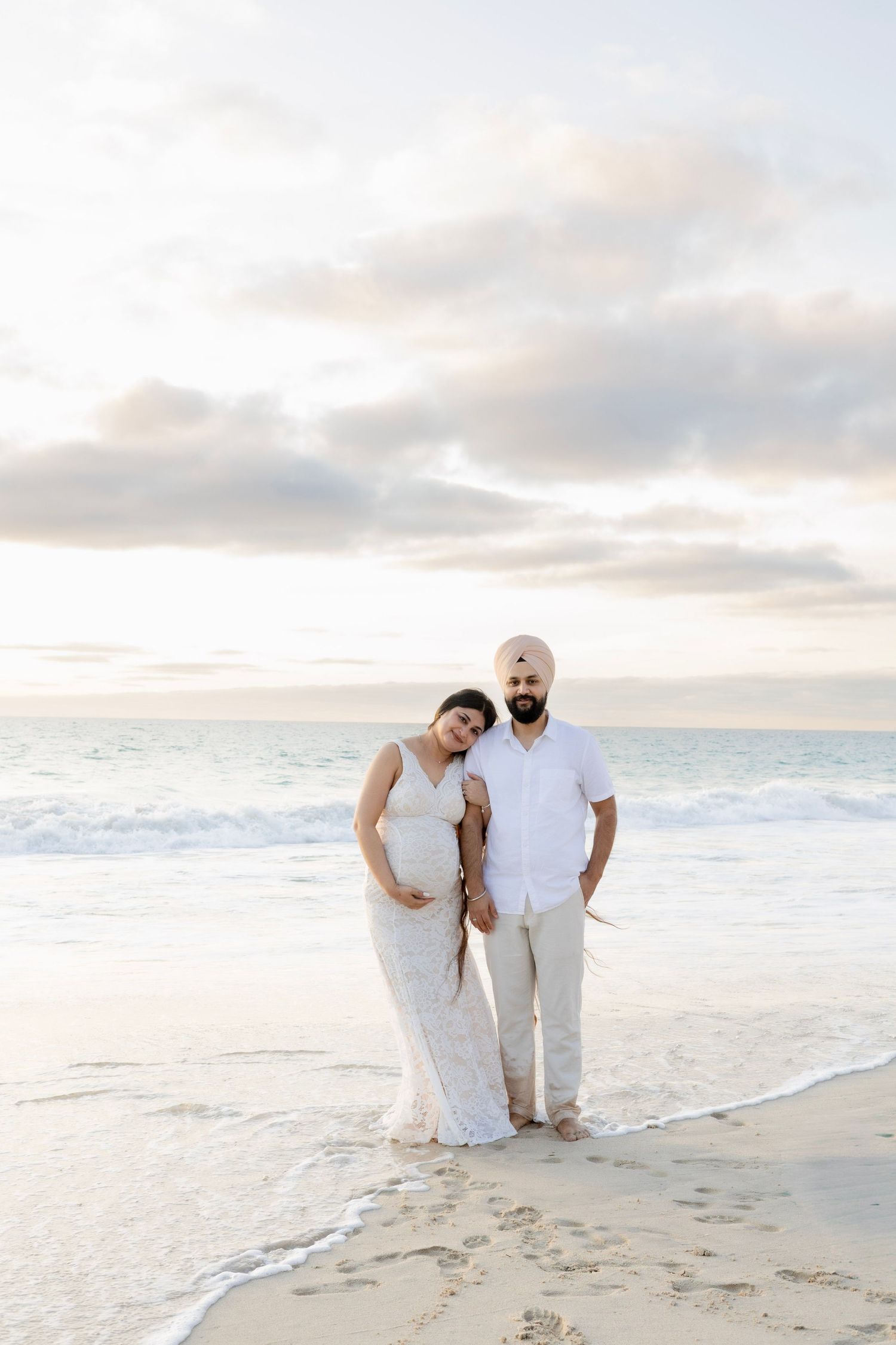 Pregnant woman in white lace dress and Sikh man standing together on beach at sunset.