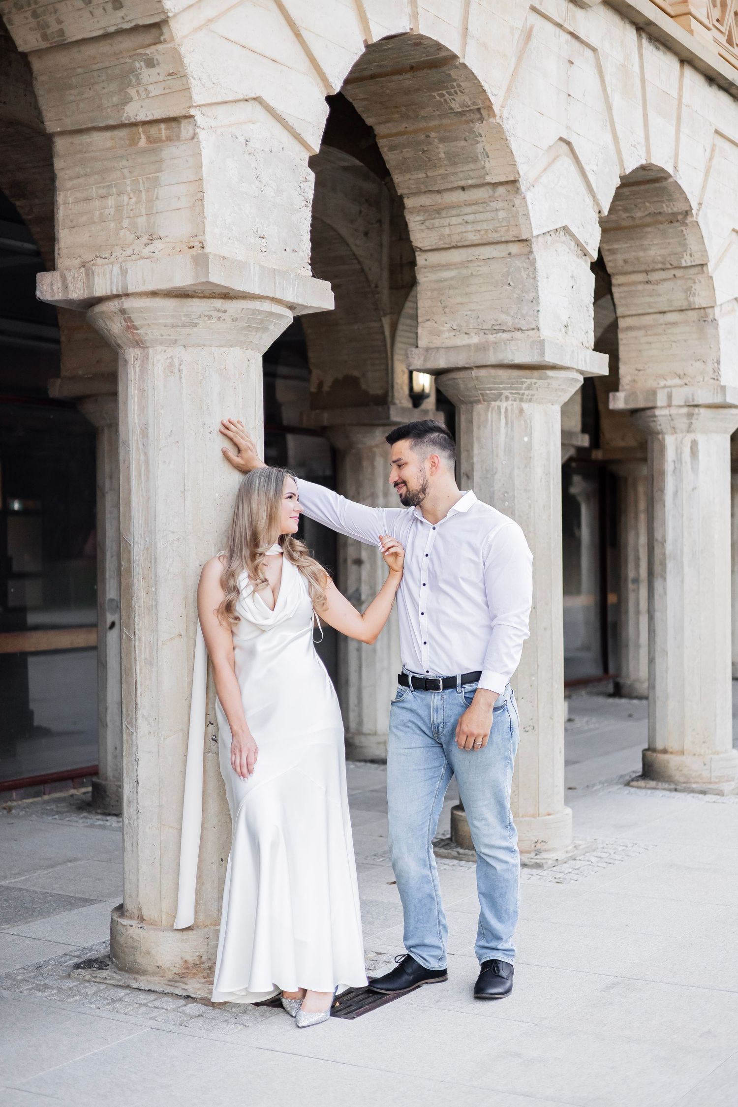 Couple dancing elegantly under stone arches, woman in white dress, man in white shirt and jeans, historic colonnade backdrop.