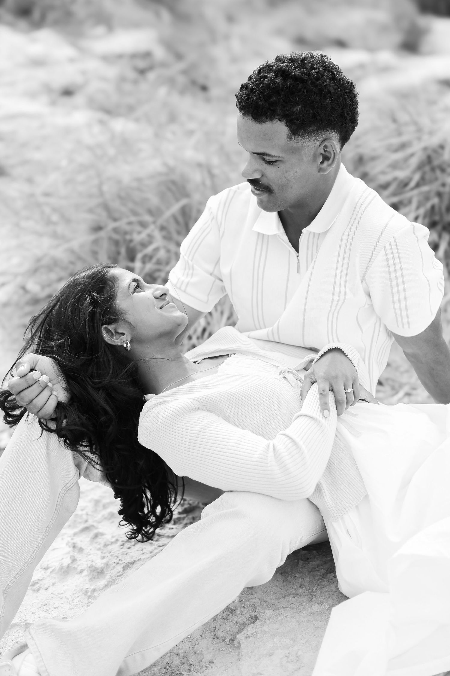 A black and white photo of a couple sharing a tender moment while sitting on beach rocks.