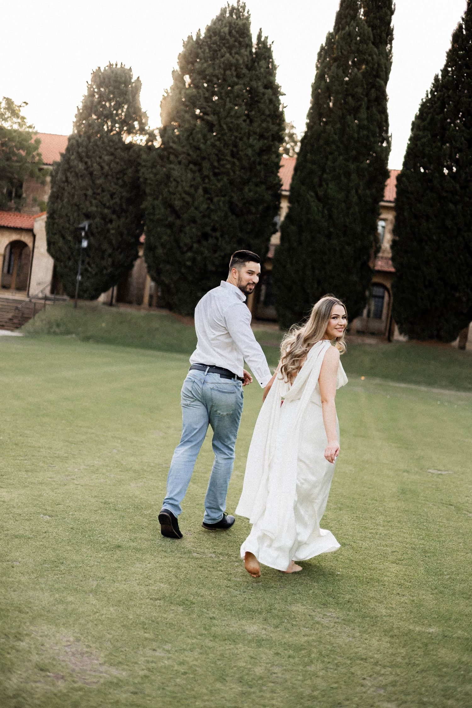 Couple walking hand in hand on lush green lawn, bride in flowing white dress smiling back at groom.