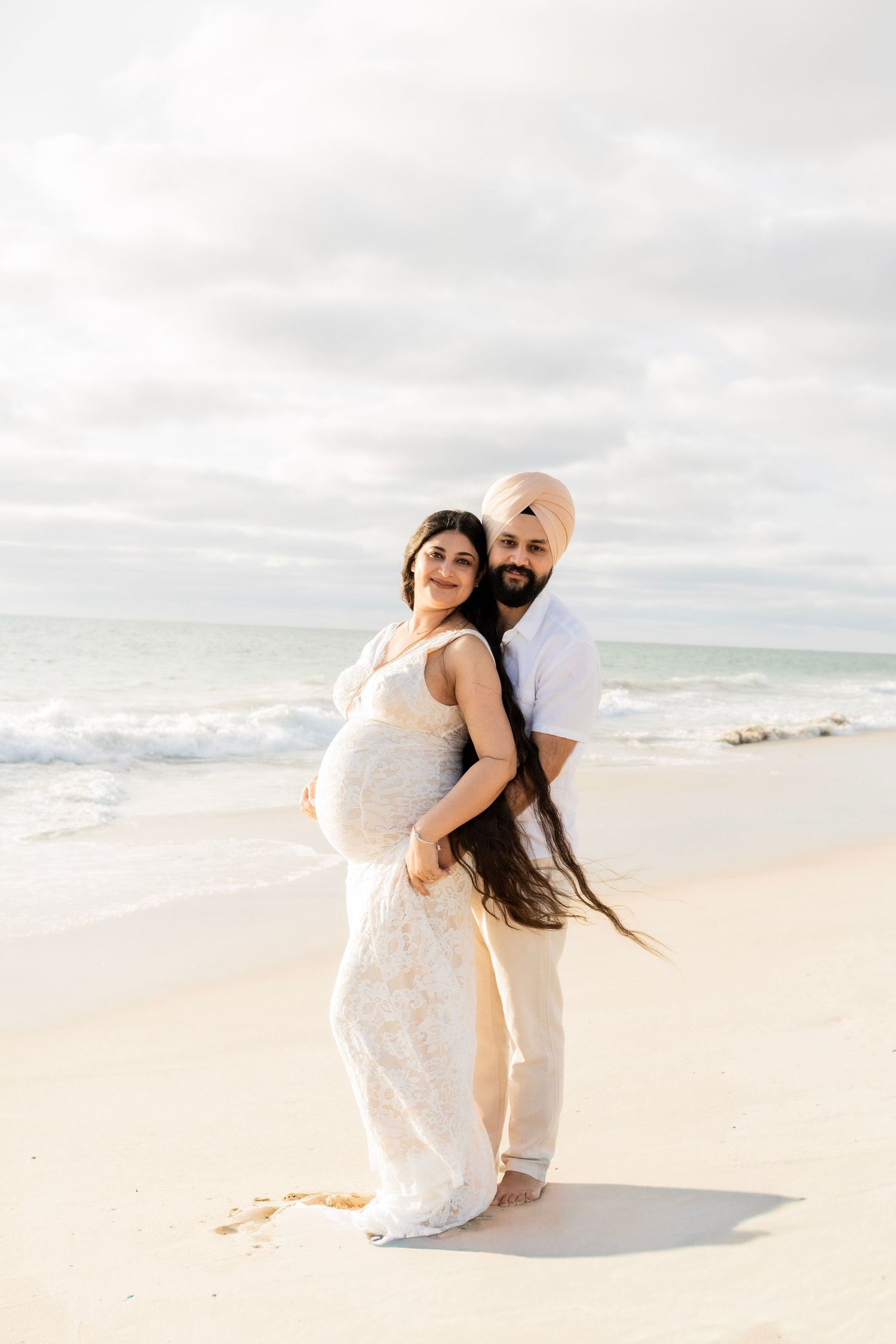 Couple posing on sunny beach, man in pink turban embracing pregnant woman in white lace gown.