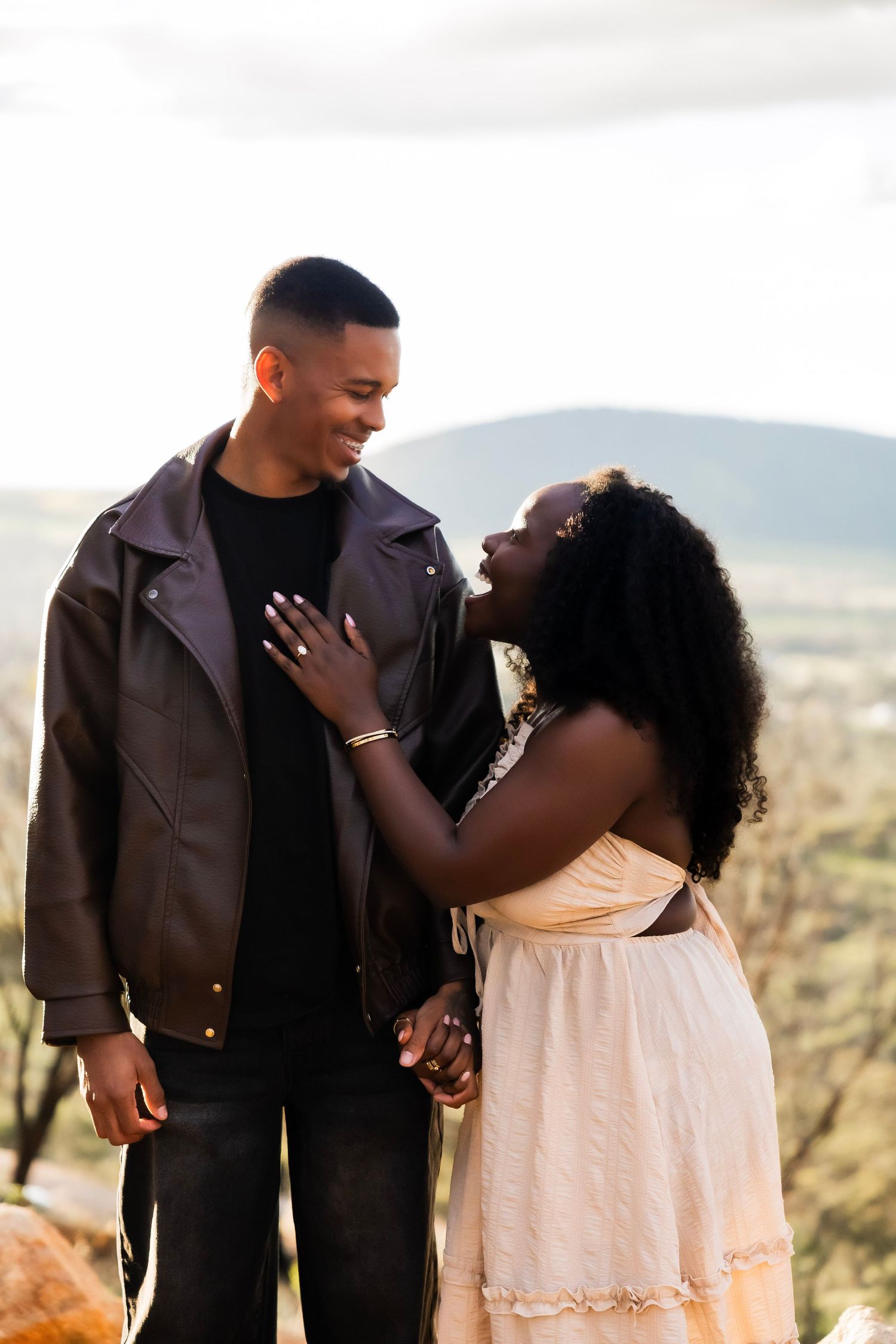 A young Black couple shares a romantic moment outdoors, holding hands with mountain views behind them.