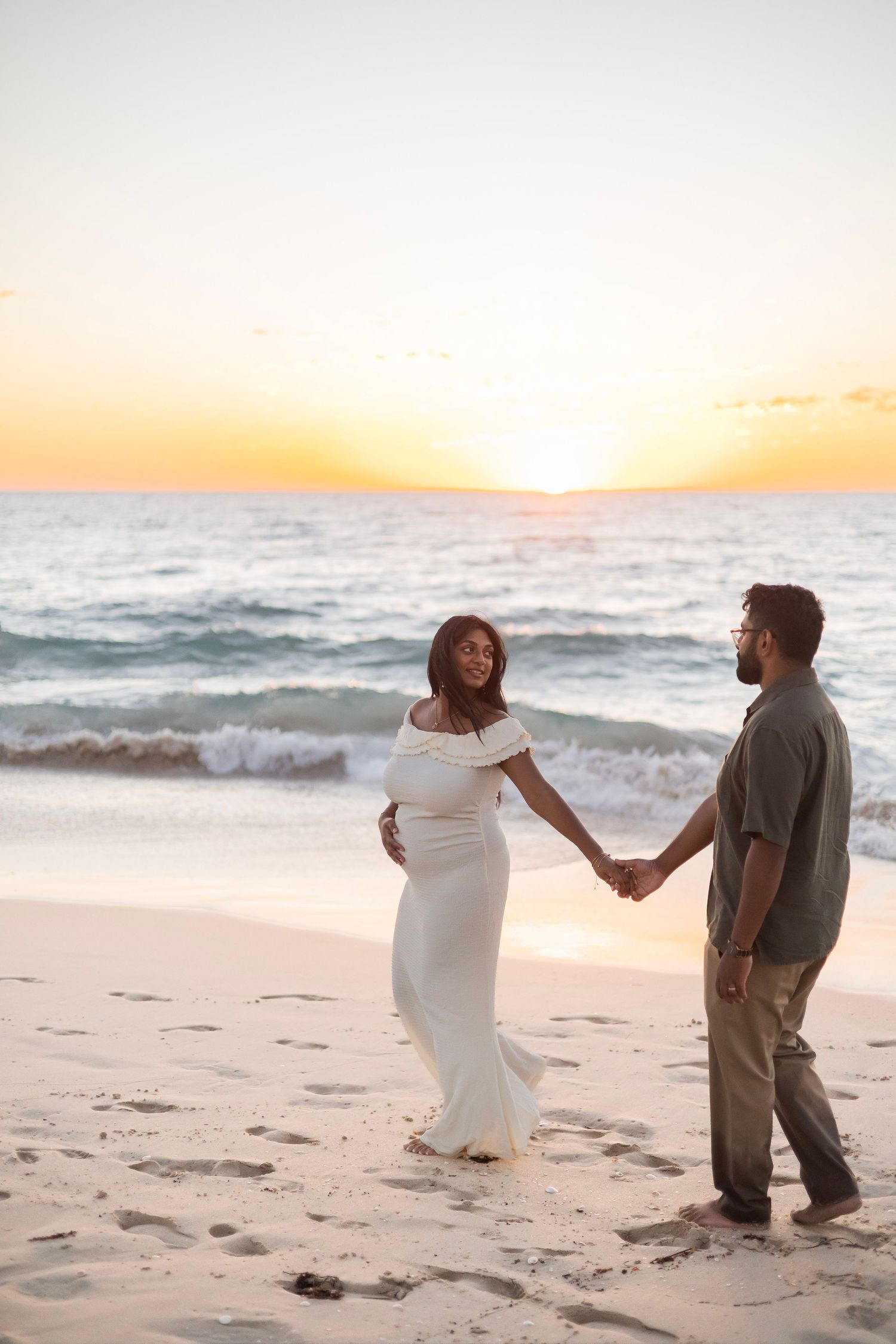 Pregnant woman in white gown holds hands with partner on beach during stunning sunset.