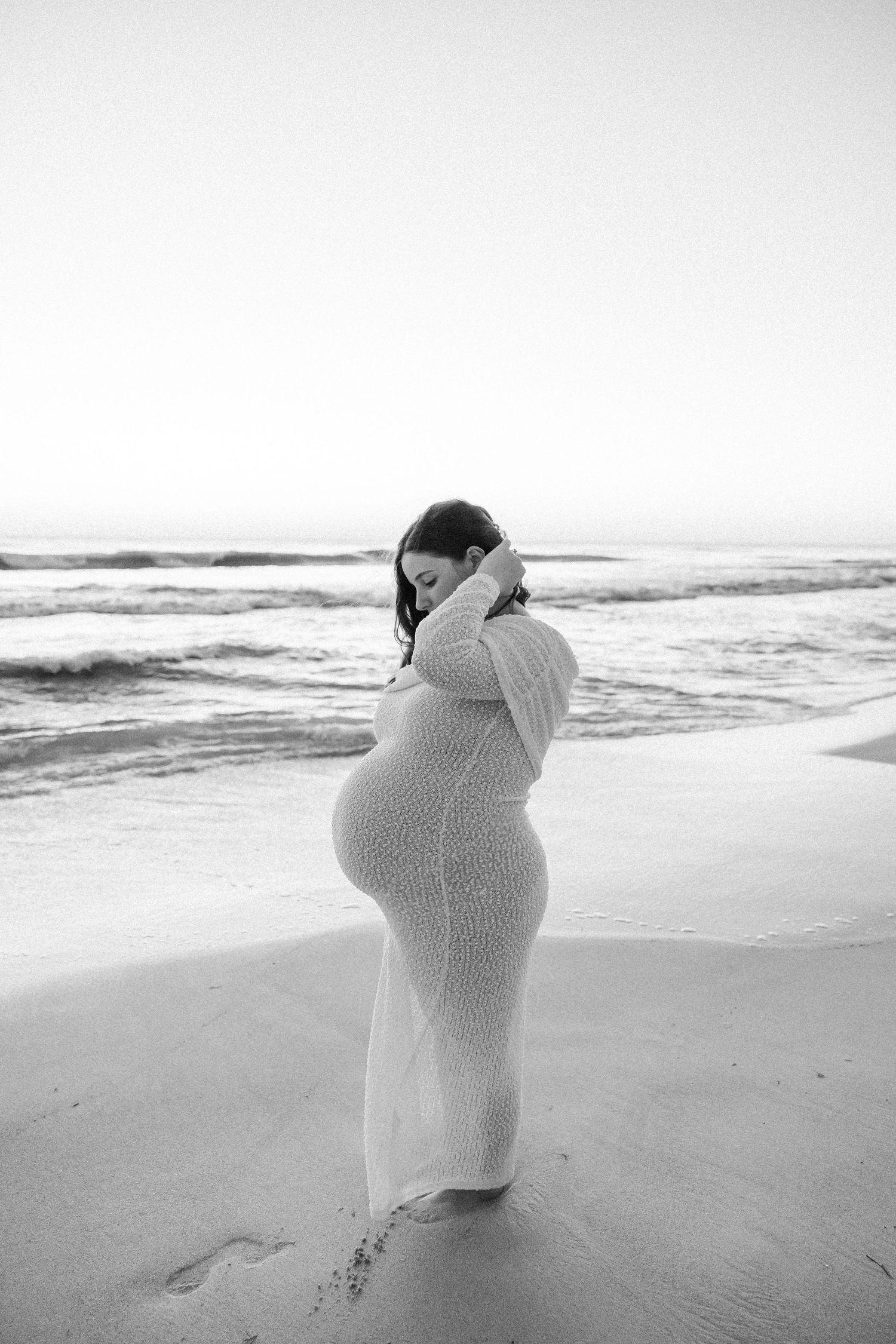 Black and white photo of pregnant woman in knit dress standing on beach, hands raised to her hair.