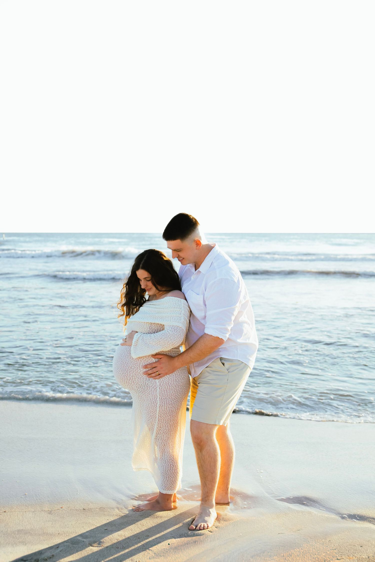 Couple sharing a tender moment on the beach, with the woman in a white maternity dress and man in white shirt.