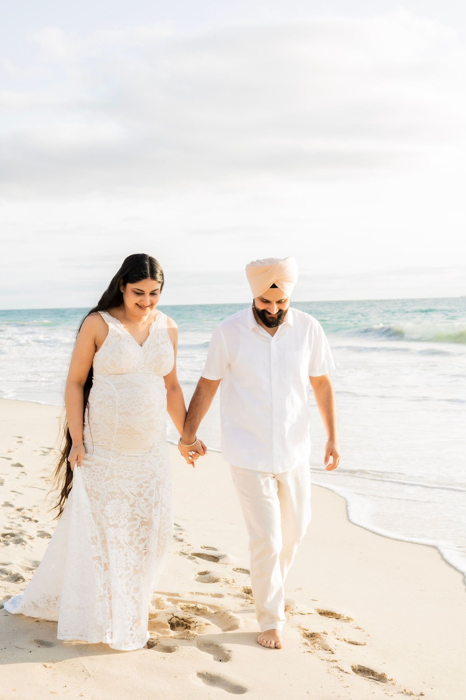 Pregnant couple walking hand in hand along ocean shoreline, woman in white lace dress, man in turban.
