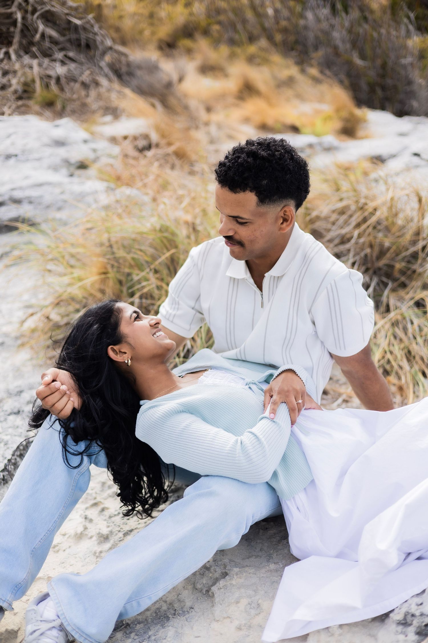 A couple shares a loving gaze while relaxing on coastal rocks surrounded by dry grass.