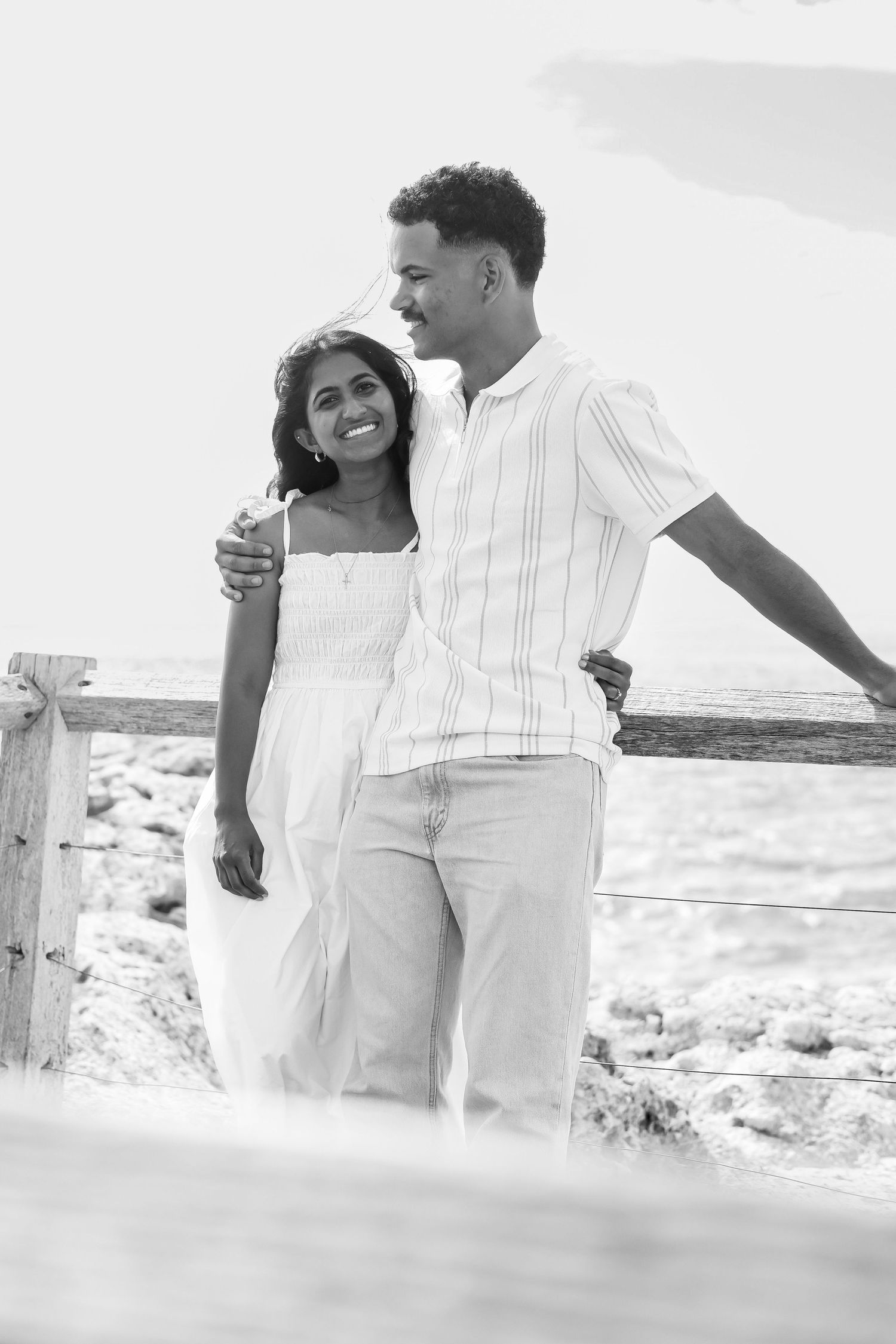 Black and white photo of a smiling couple embracing by a wooden fence railing near the ocean.