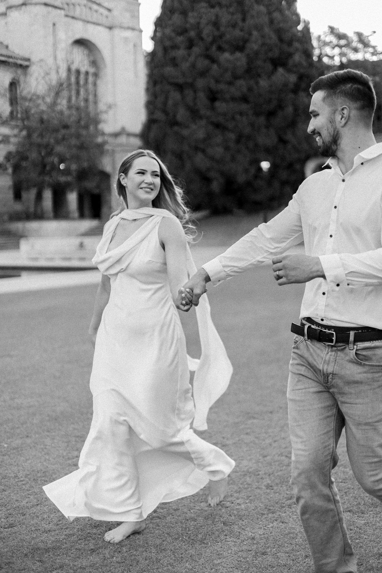 Black and white photo of smiling bride in white dress holding hands with groom while walking on green lawn.