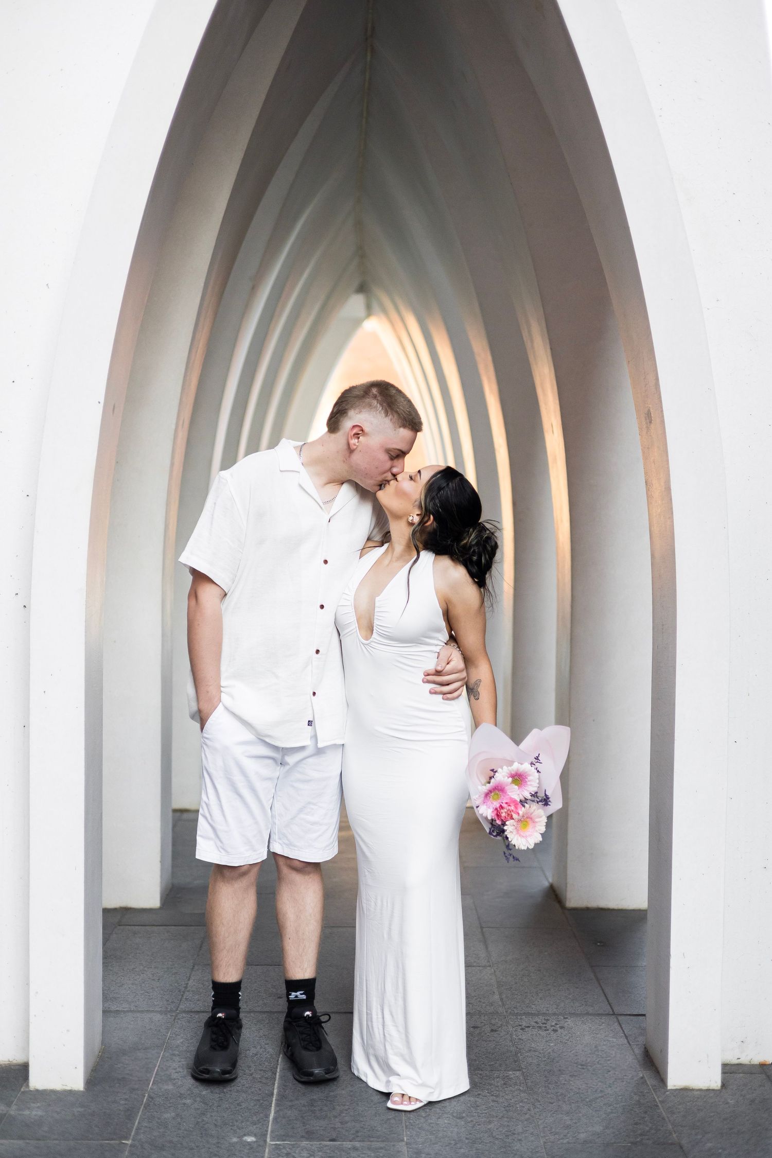 Couple sharing a romantic kiss in a white arched tunnel, woman in white gown holding a pink bouquet.