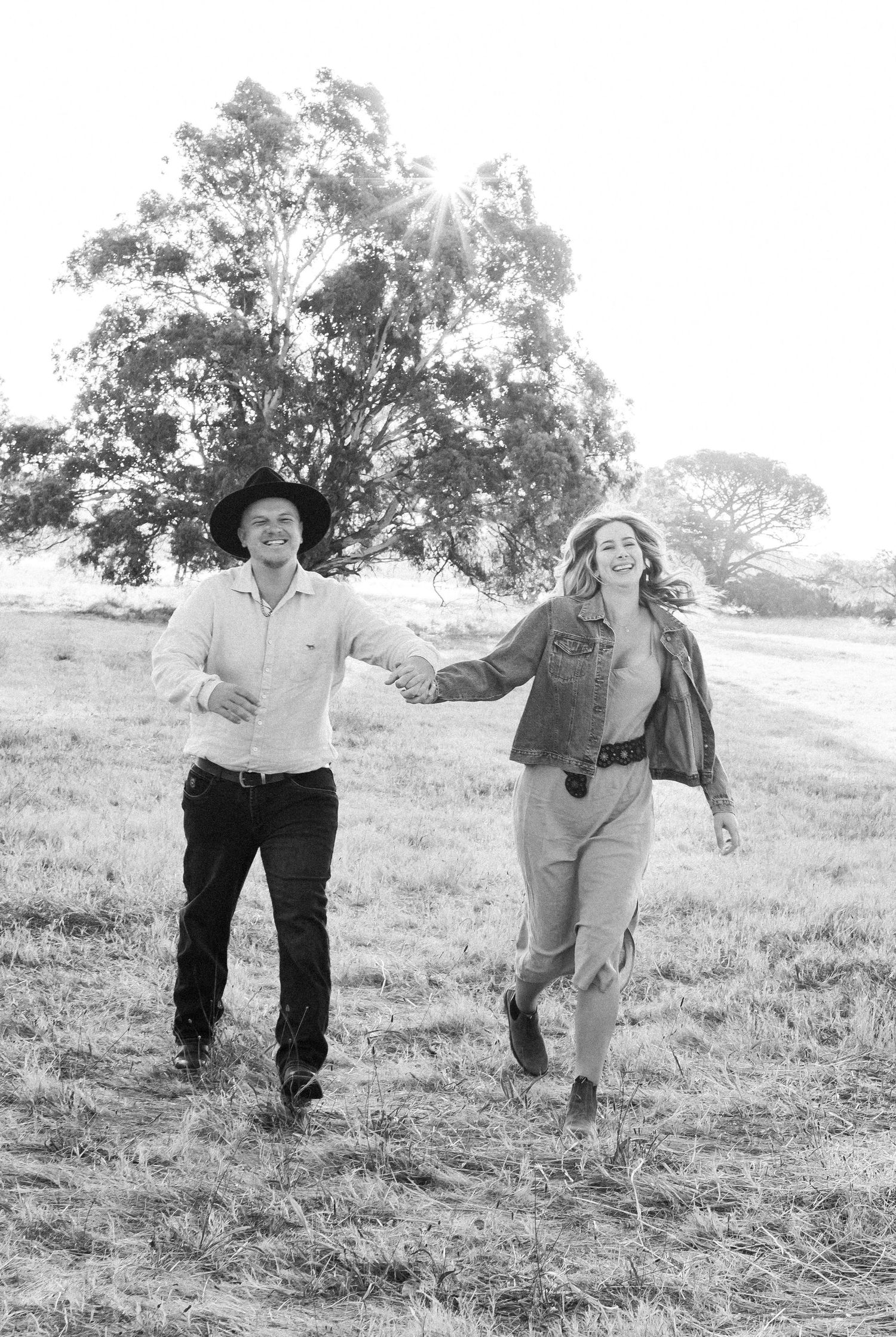 Black and white photo of a joyful couple holding hands and running through a sunlit open field together.