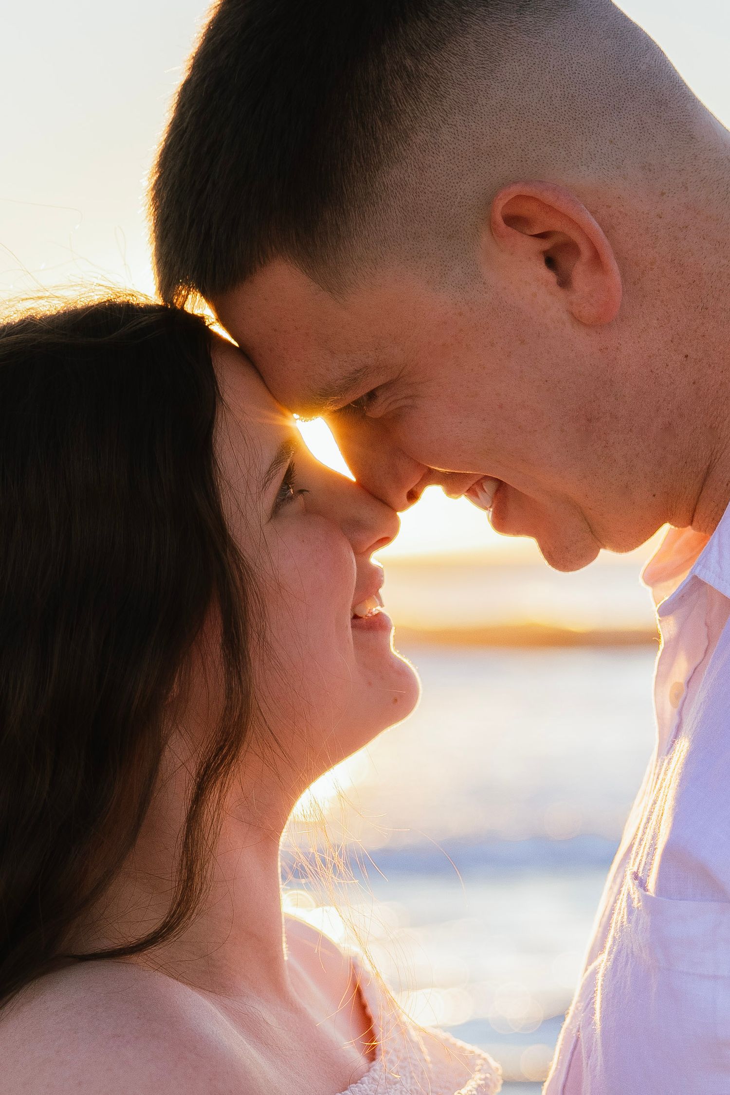 Romantic couple touching foreheads at sunset on a beach, sharing an intimate moment with golden backlight.