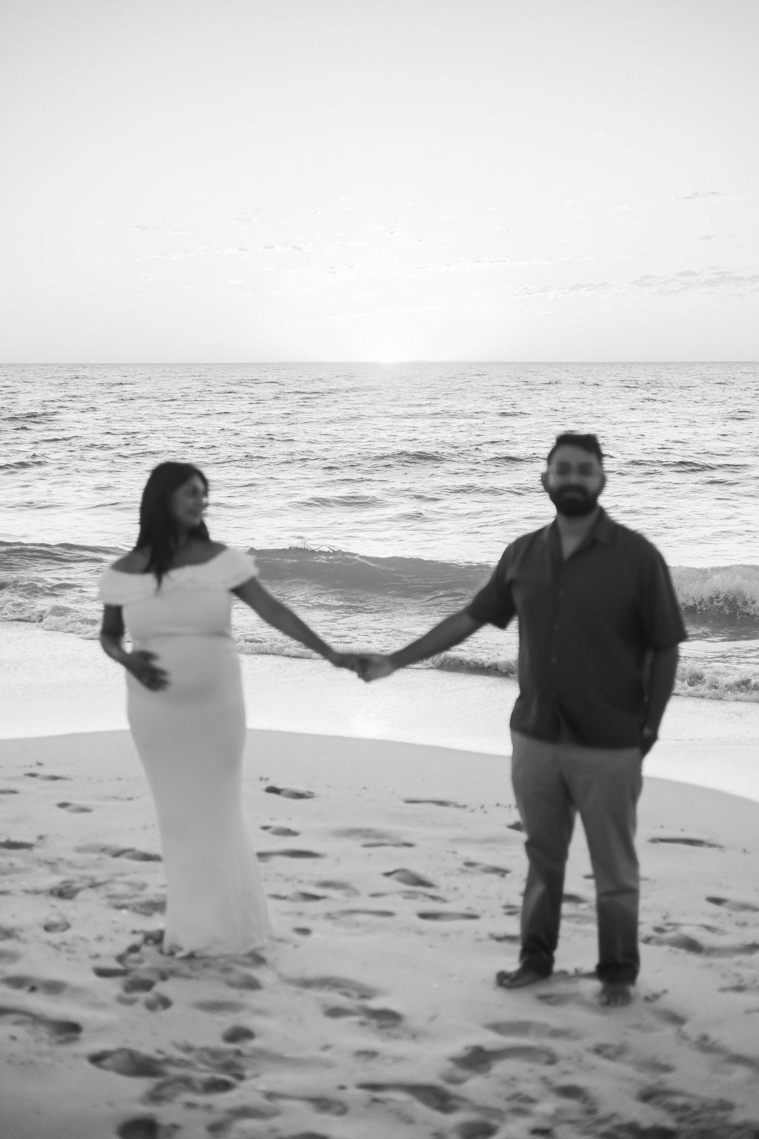 Black and white photo of pregnant couple holding hands on beach at sunset.