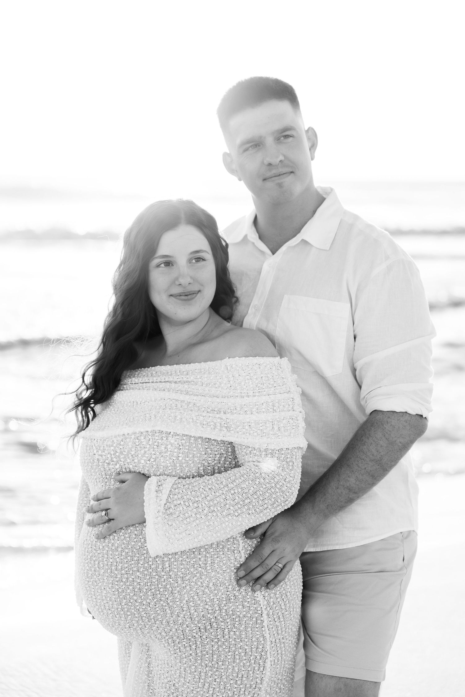 Black and white beach maternity photo of a couple, pregnant woman in off-shoulder dress, man embracing her from behind.