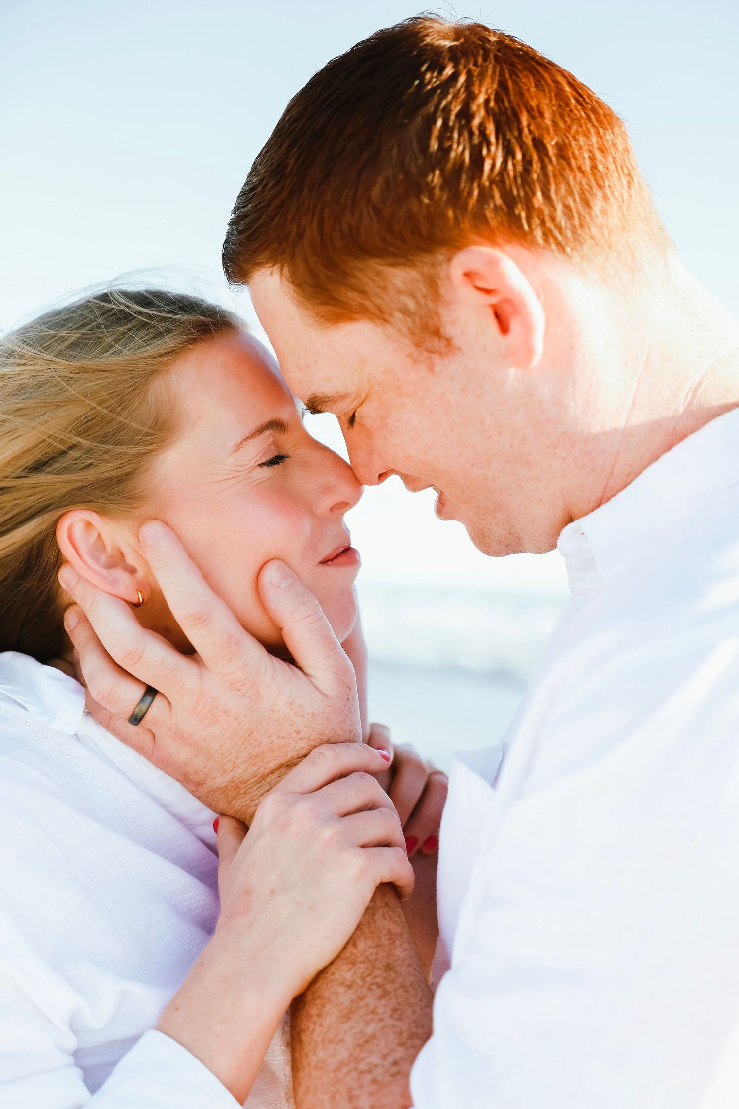 Romantic close-up of couple almost kissing on beach, man with red hair in white shirt, woman with blonde hair.