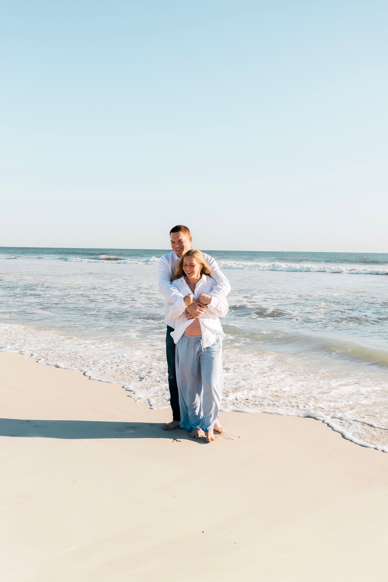 Couple embracing on a white sand beach with ocean waves, man in white shirt hugging woman from behind near shoreline.