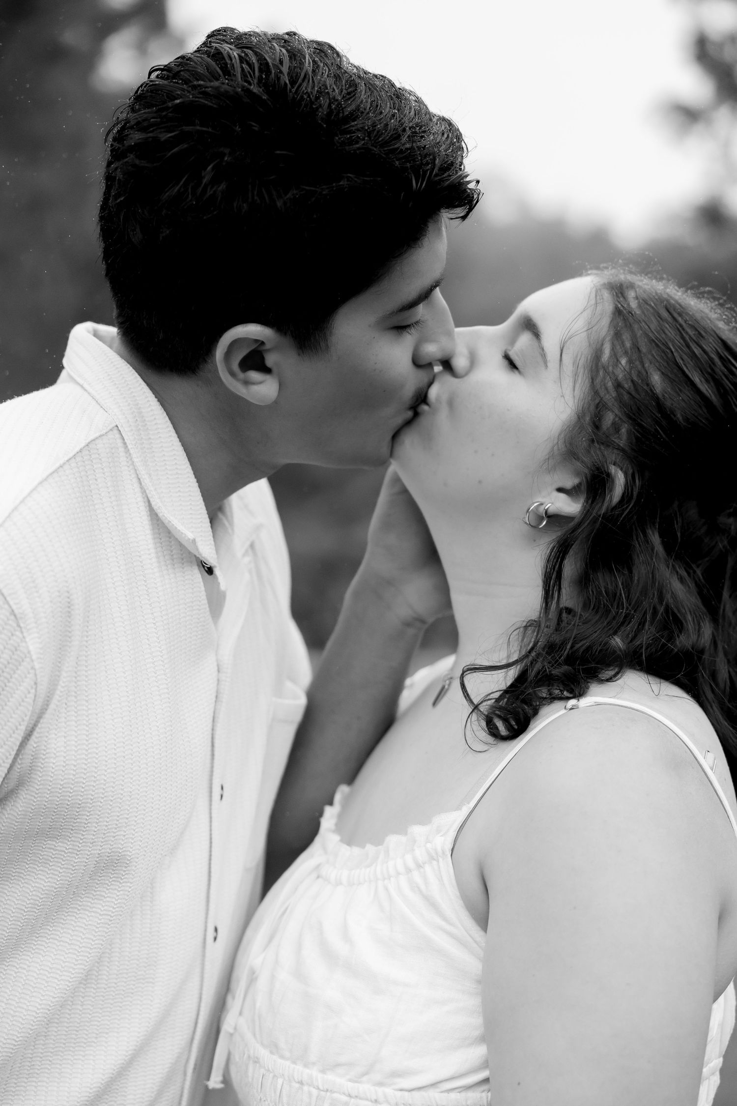 Black and white photo of a young couple sharing a romantic kiss outdoors, both dressed in white.