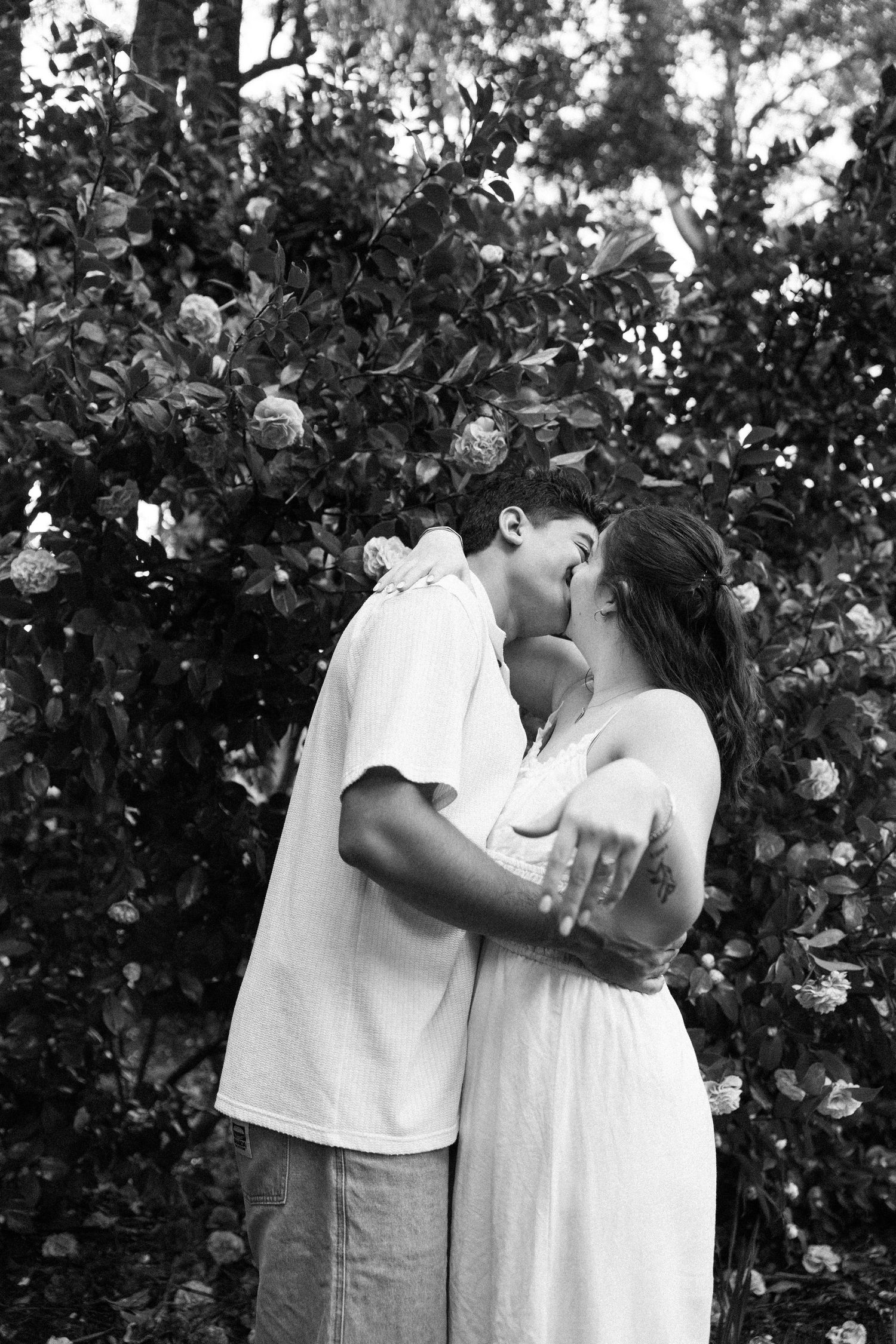 Black and white photo of a couple kissing in front of a flowering bush in a lush garden setting.