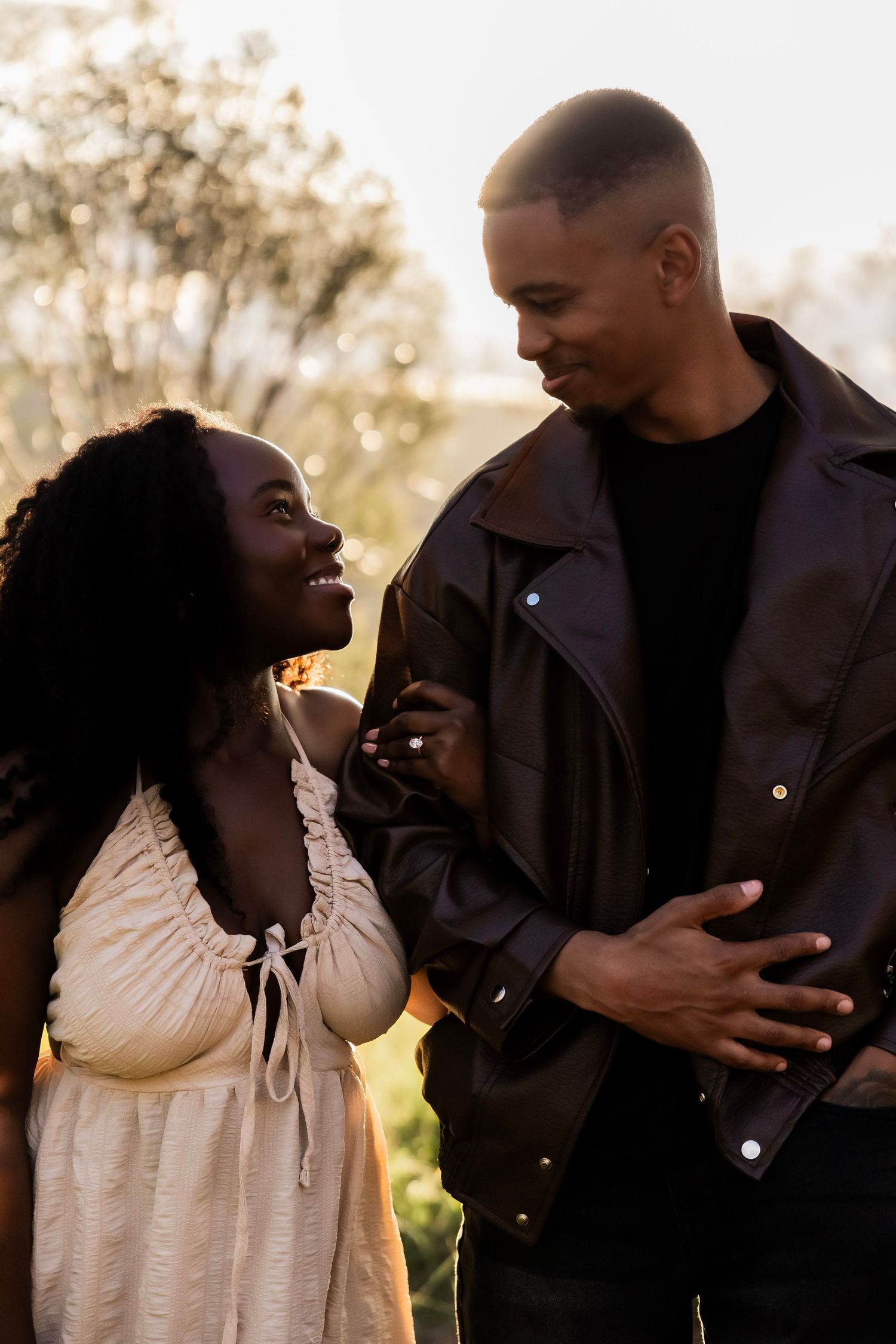 A smiling Black couple shares a tender moment outdoors at golden hour, woman in white dress, man in black leather jacket.