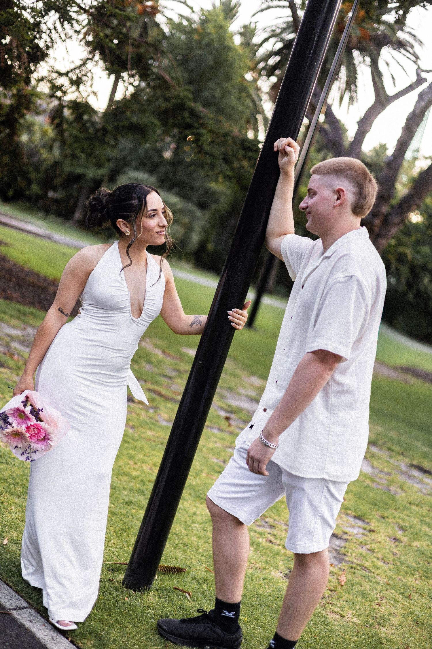 Bride and groom in white sharing a playful moment by black pole in park.