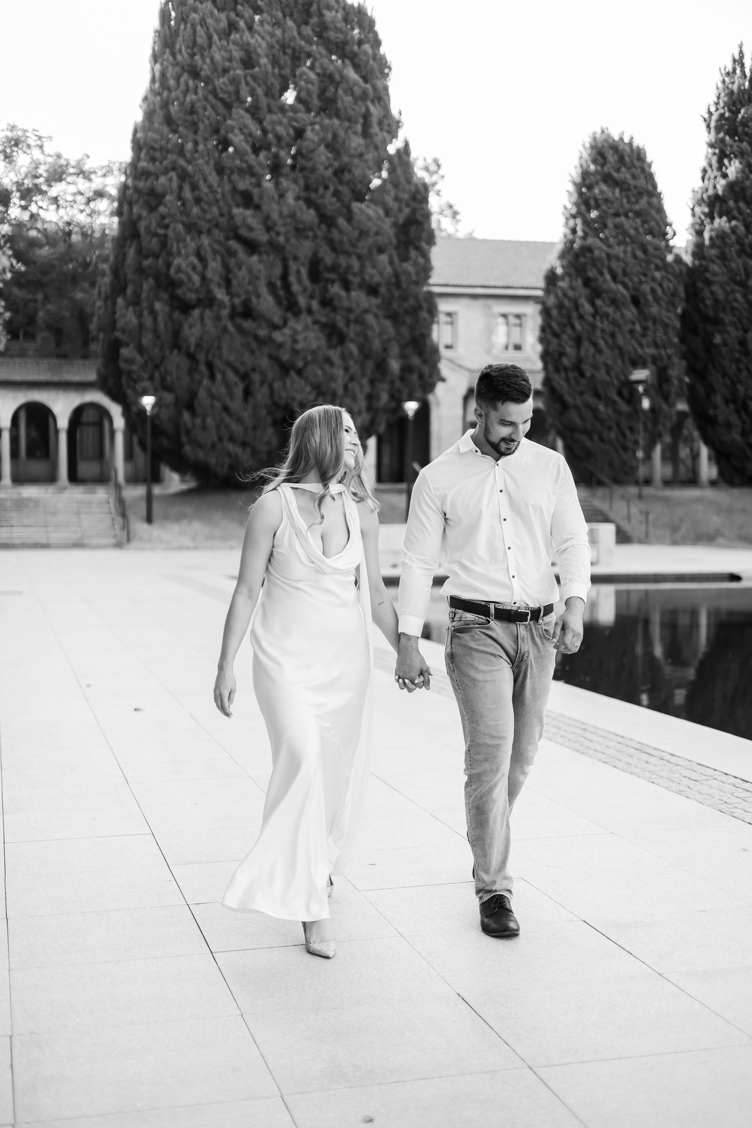 Black and white photo of couple holding hands strolling near reflecting pool with cypress trees behind.