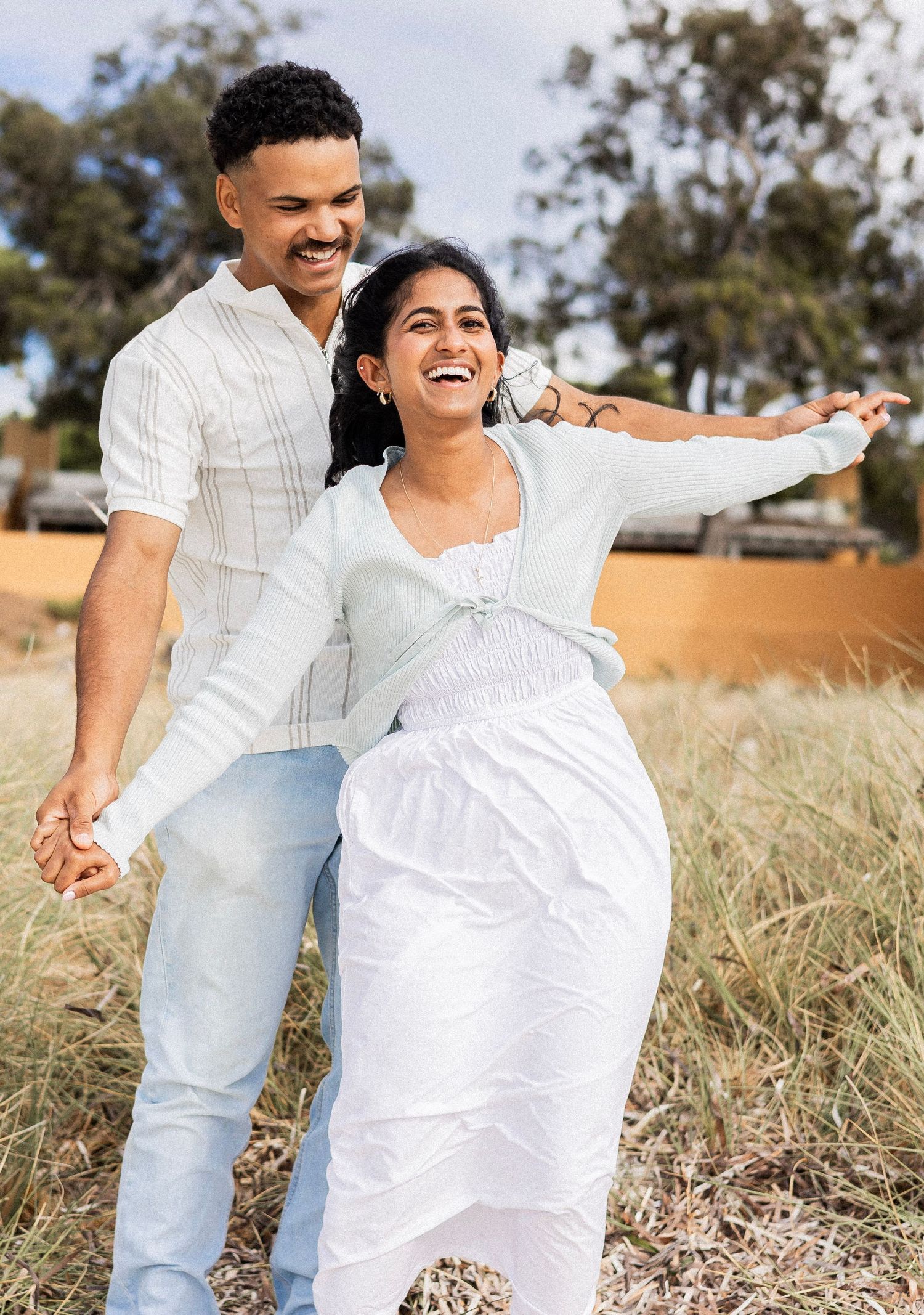 Joyful couple in white outfits laughing and spinning together in a sunny outdoor field.