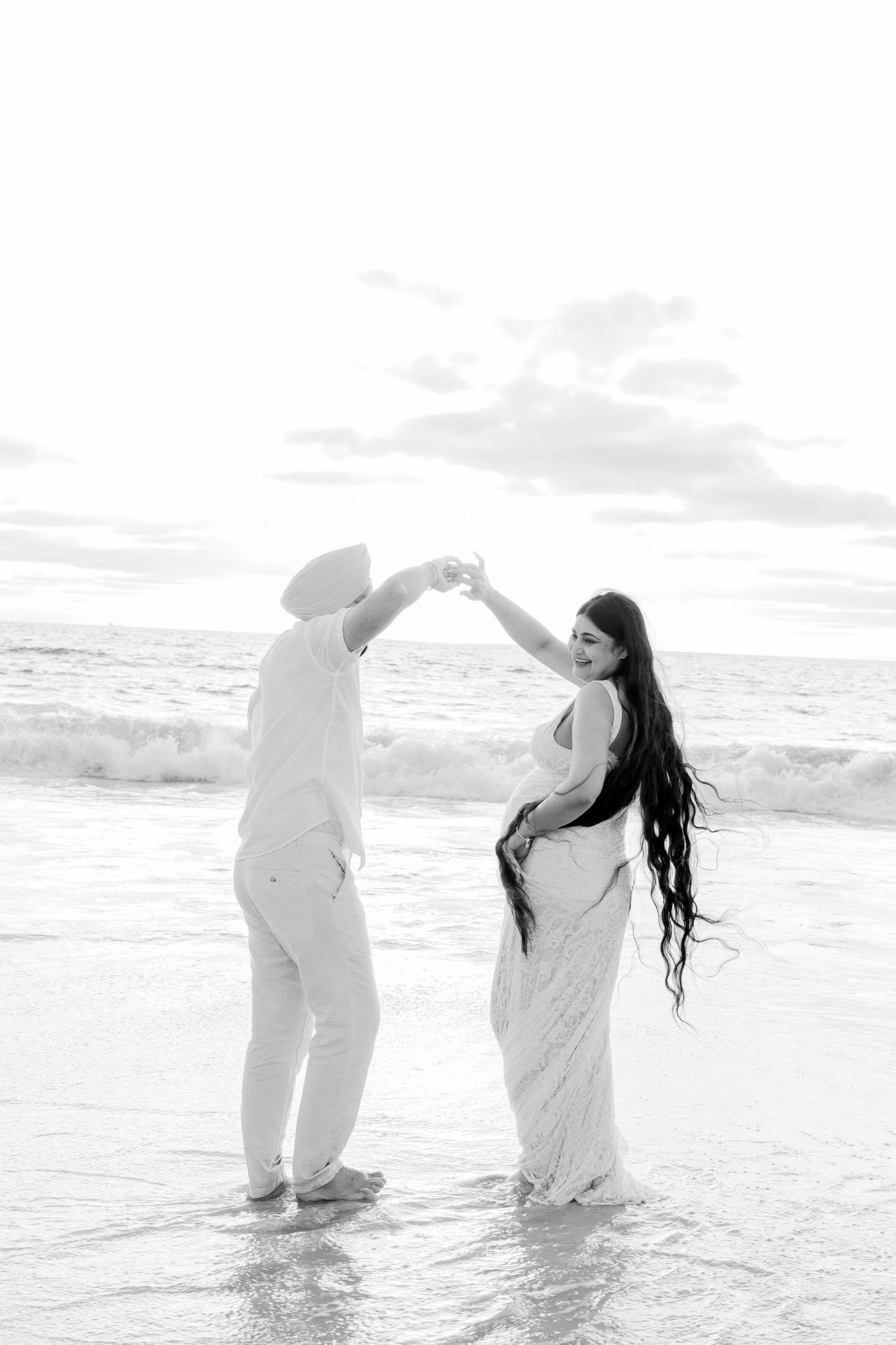 Black and white photo of a couple dancing barefoot in ocean waves on a beach at sunset.