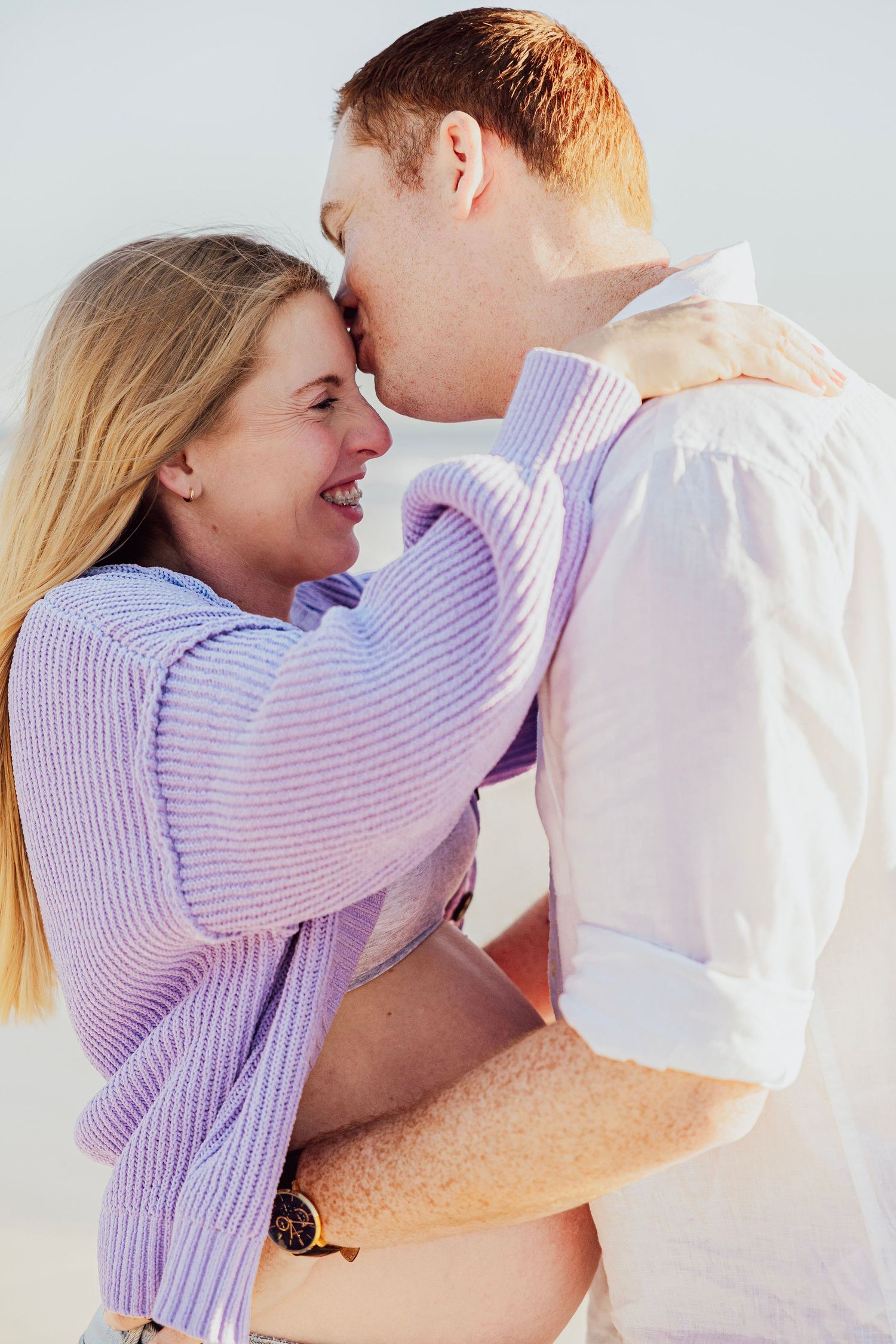 A smiling couple in pastel sweaters share a tender nose-to-nose moment on a beach, embracing warmly.