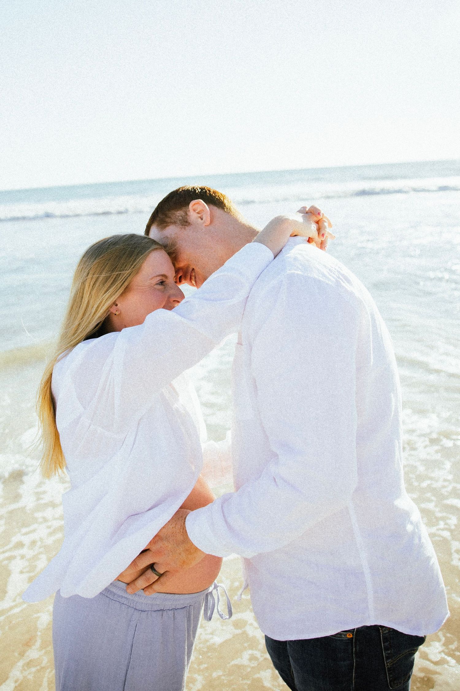 Happy pregnant couple in matching white shirts embracing and dancing together on a sunny beach shoreline.