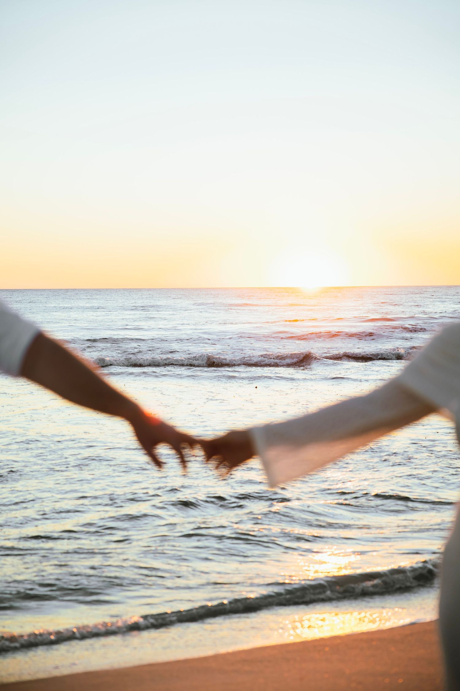 Close-up of couple holding hands on beach at sunset with golden sky and ocean waves behind them.