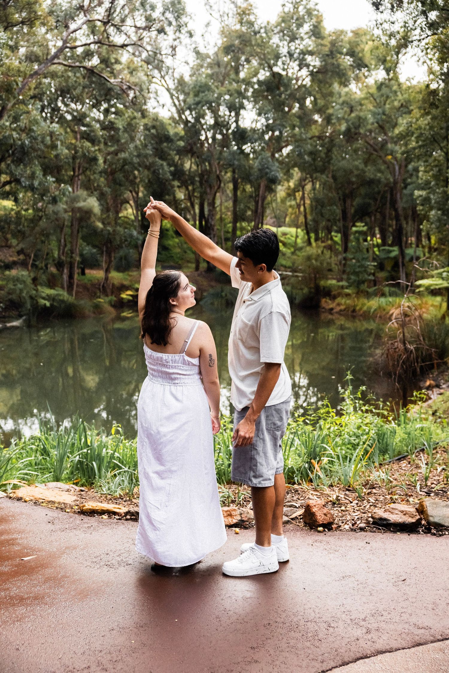 Couple dancing together near a tranquil pond surrounded by lush green trees and plants.