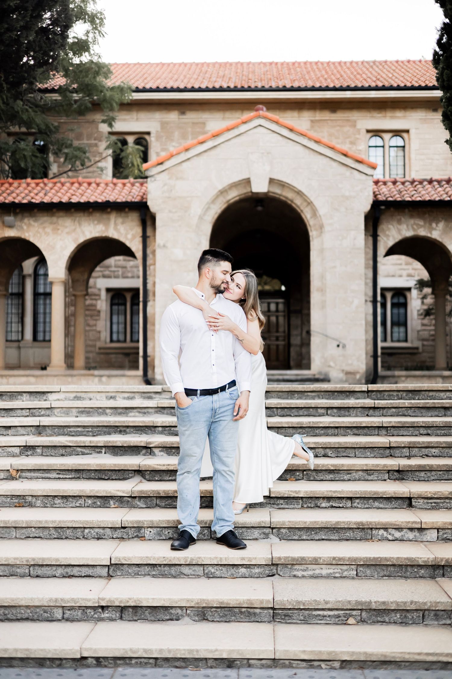 Couple embracing on stone steps before a historic building with arched doorways and red tile roof.