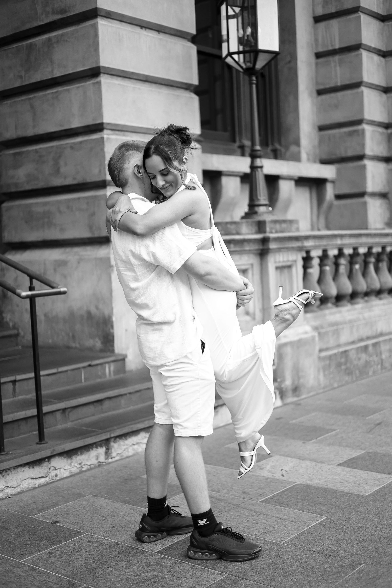 Black and white photo of couple embracing joyfully outside classic building.